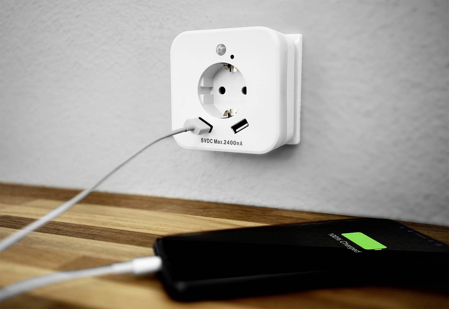 Smartphone mit Ladeanzeige liegt auf Tisch, verbunden mit einer Steckdose über ein USB-Kabel. Steckdose hat zusätzliche USB-Ports.
