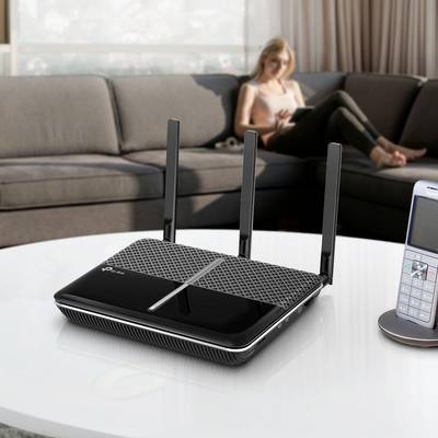 Ein WLAN-Router mit drei Antennen steht auf einem Tisch im Wohnzimmer; im Hintergrund sitzt eine Person auf einem Sofa.