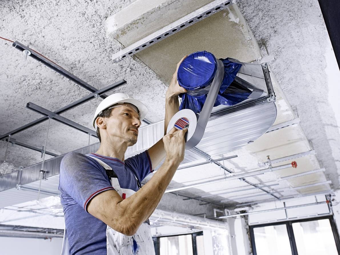 Ein Arbeiter mit Bauhelm versiegelt eine Lüftungsöffnung an der Decke in einem Rohbau mit Klebeband, um sie abzudichten.