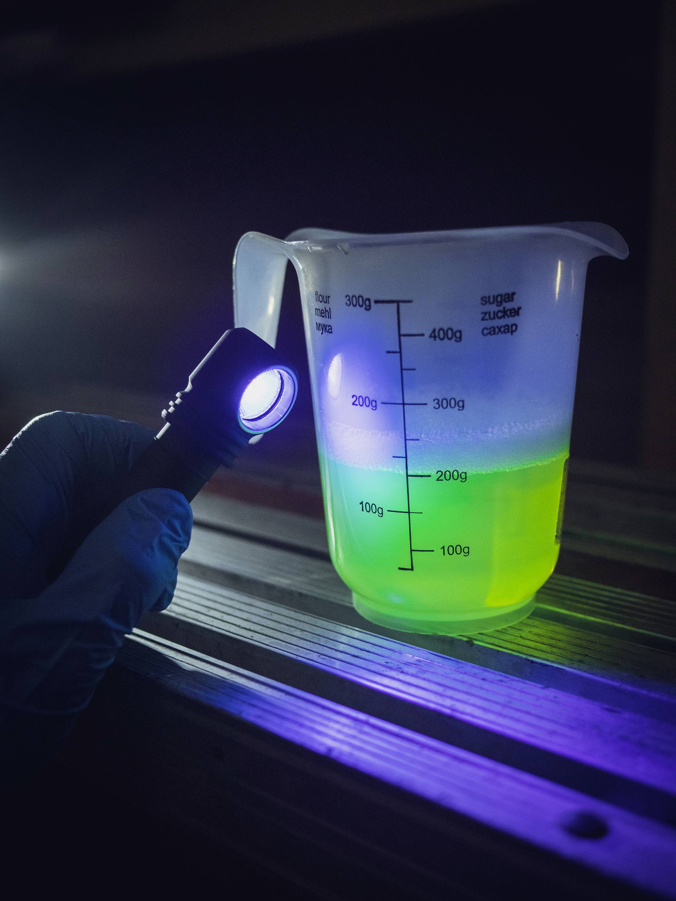 Ein Messbecher mit grün leuchtender Flüssigkeit neben einer Hand mit Taschenlampe auf einem Tisch.