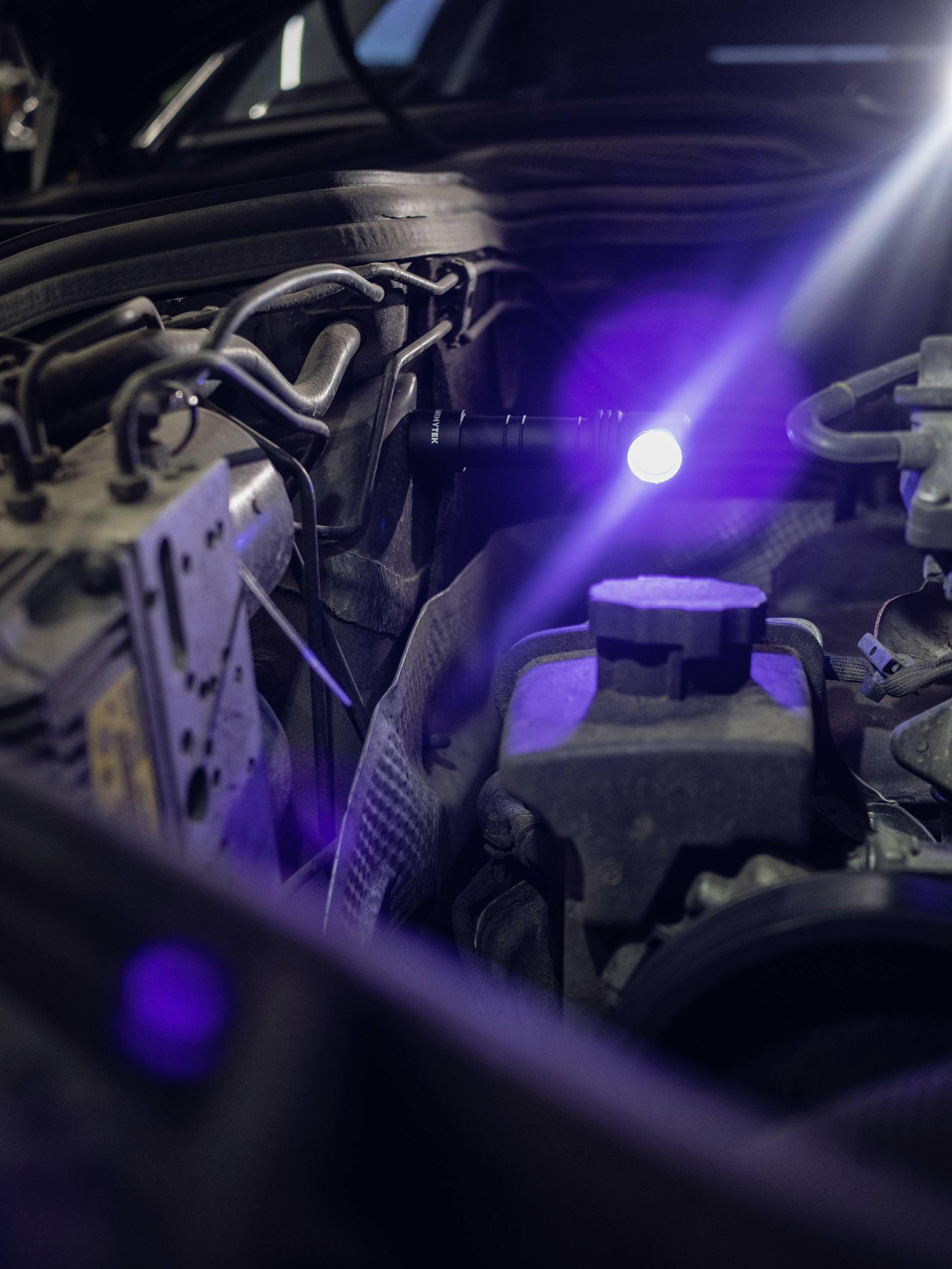 Motorraum eines Autos bei geöffneter Motorhaube, beleuchtet von einer Taschenlampe. Verschiedene Autoteile sind sichtbar.