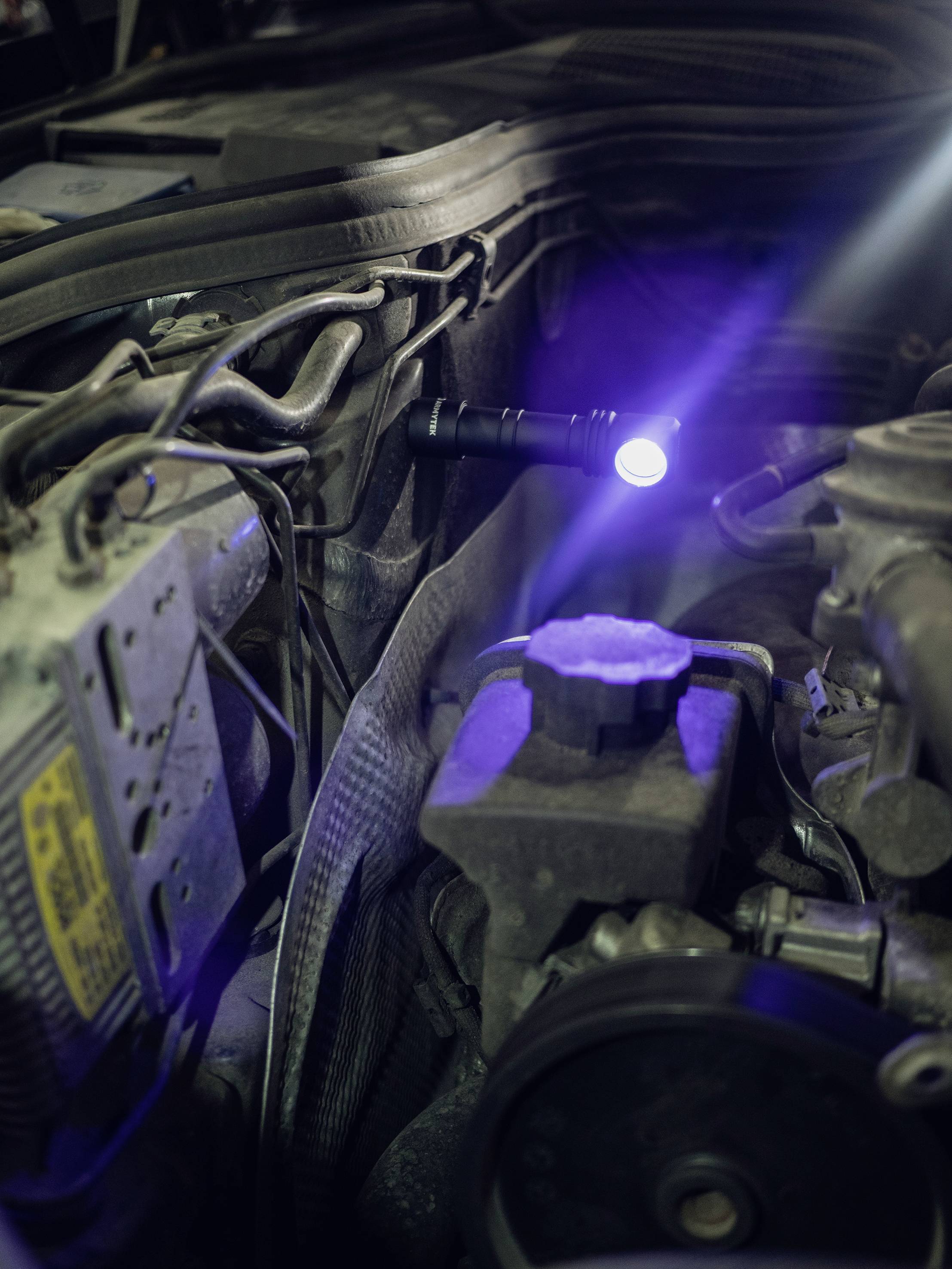 Eine Taschenlampe leuchtet im Motorraum eines Fahrzeugs, umgeben von Kabeln und mechanischen Teilen.