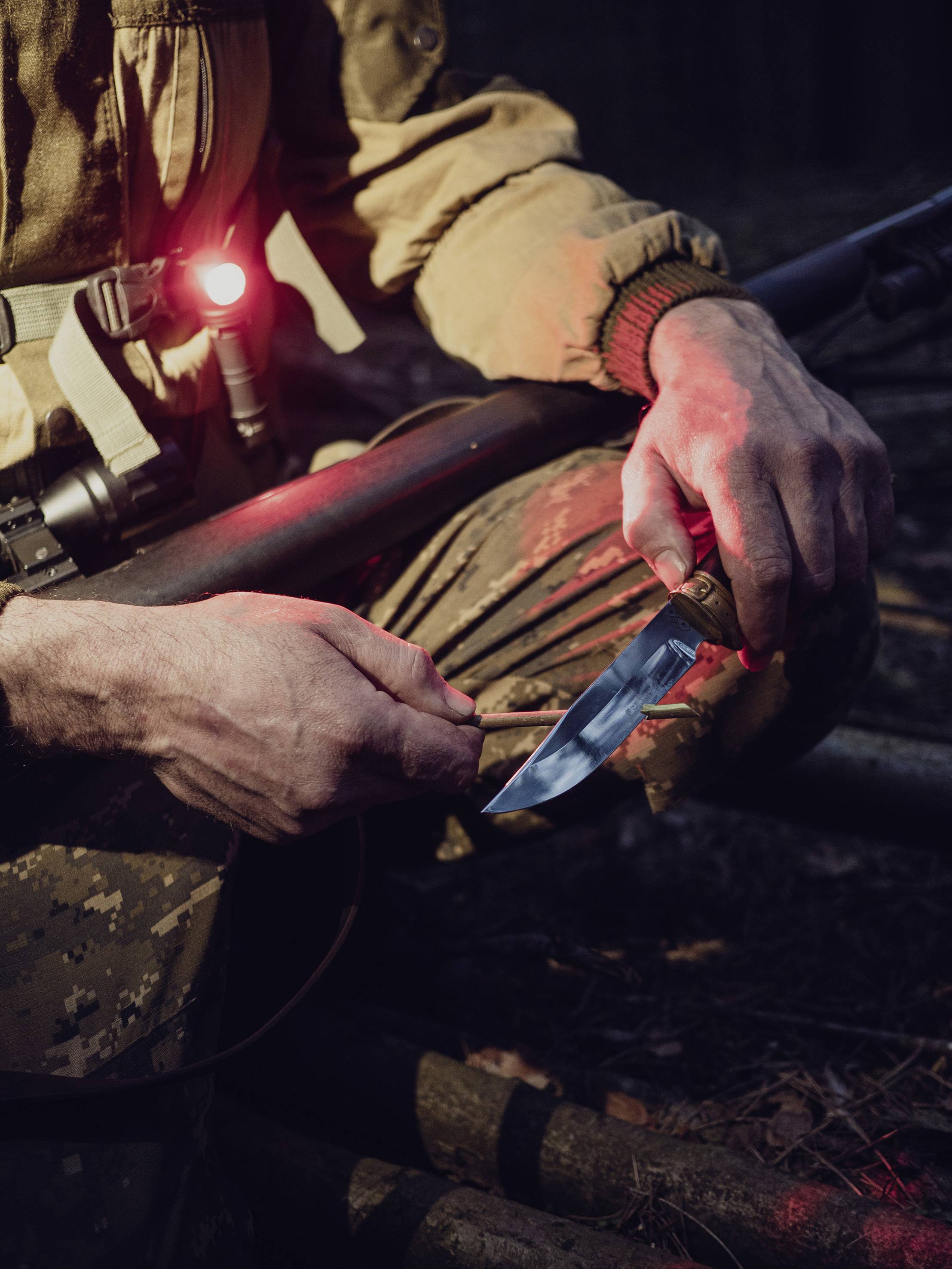 Eine Person in Tarnkleidung hockt im Wald und schärft ein Messer, gehalten neben einem Gewehr. Eine rote Lampe leuchtet an ihrer Kleidung.
