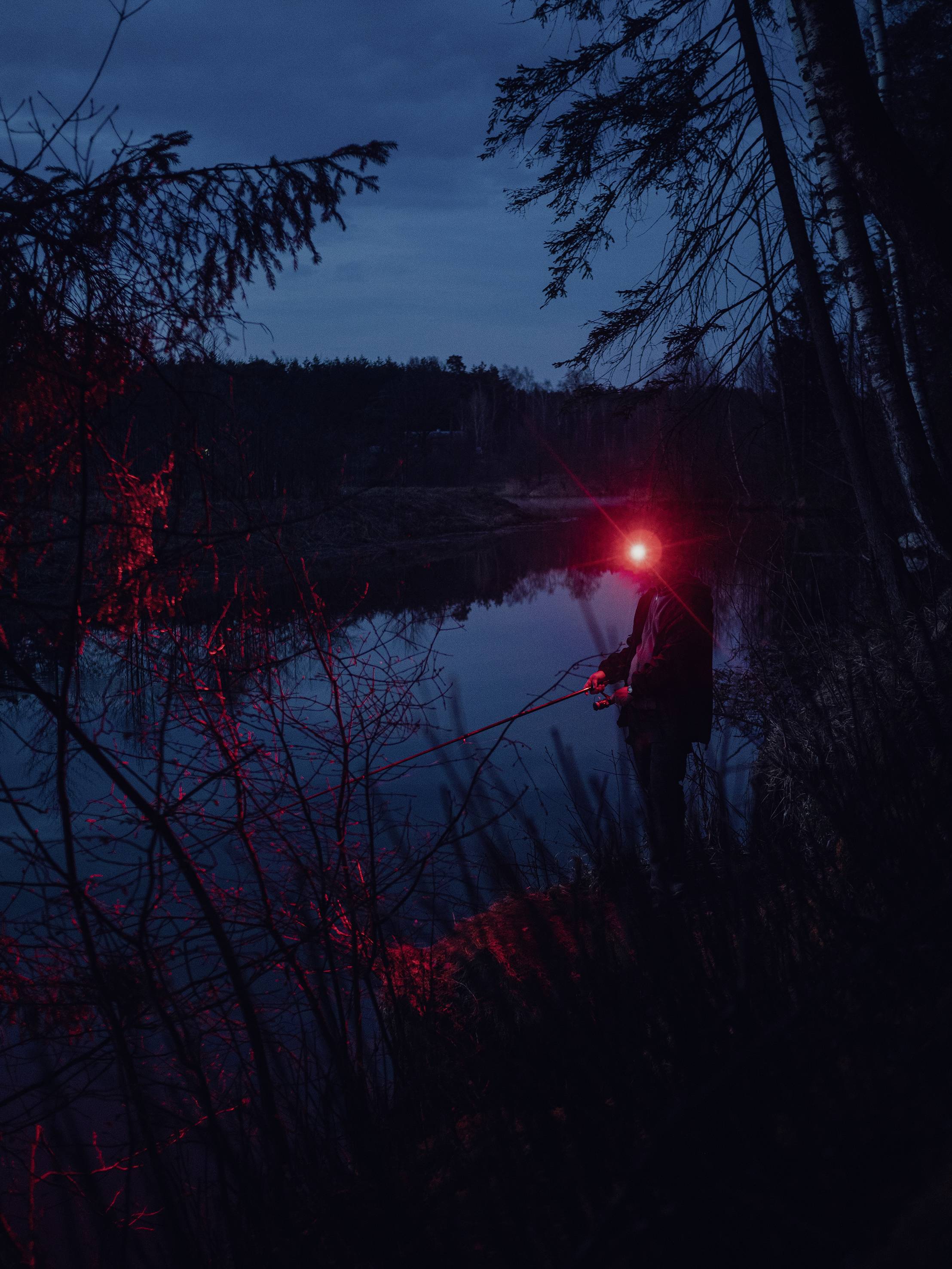 Eine Person angelt nachts am Ufer eines Sees. Sie trägt eine Stirnlampe, die rotes Licht ausstrahlt. Die Umgebung ist von Bäumen umgeben.