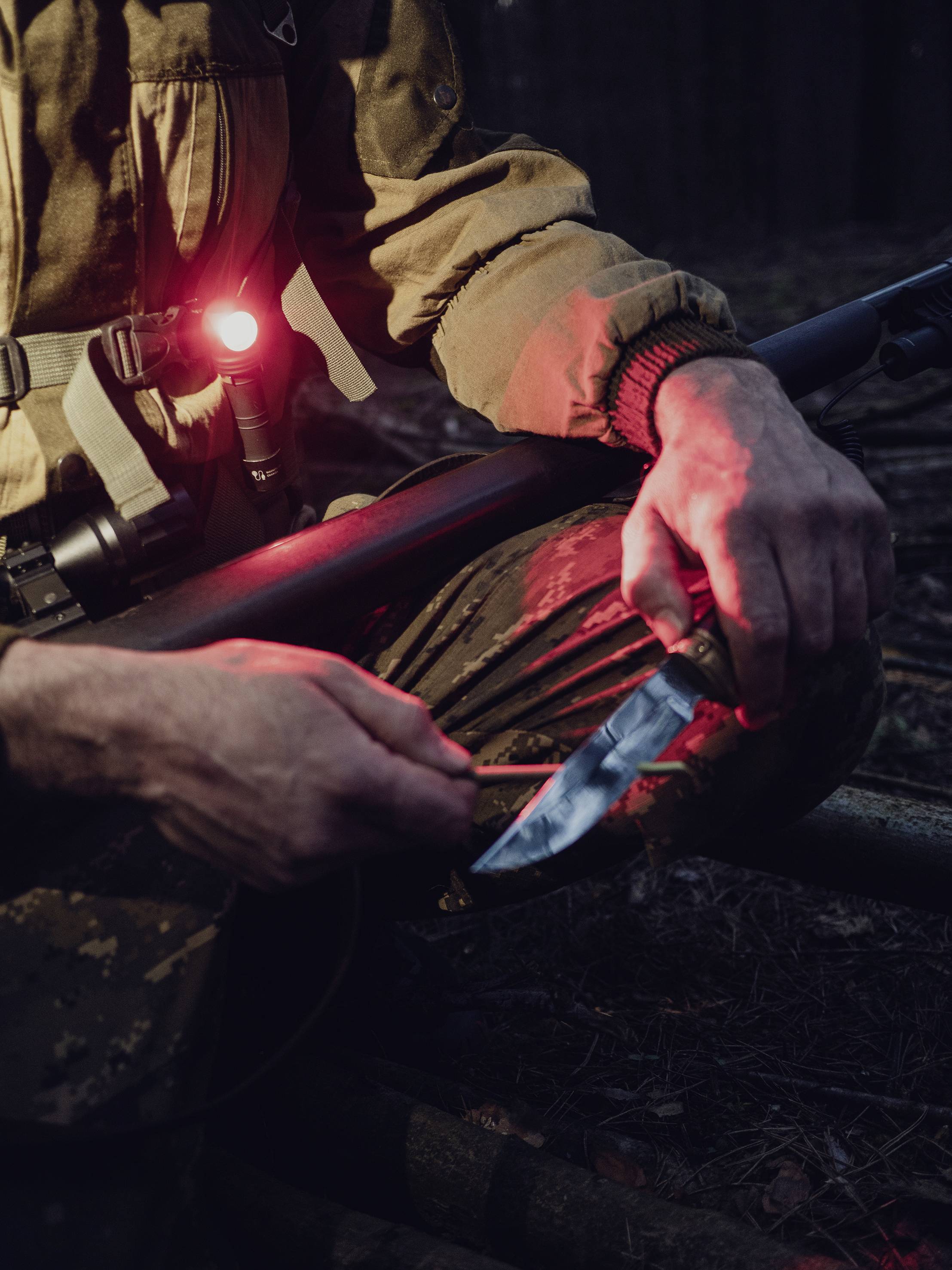 Eine Person im Tarnanzug, beleuchtet von einer roten Lampe, hält ein Gewehr und ein Messer im Wald bei Dämmerung.