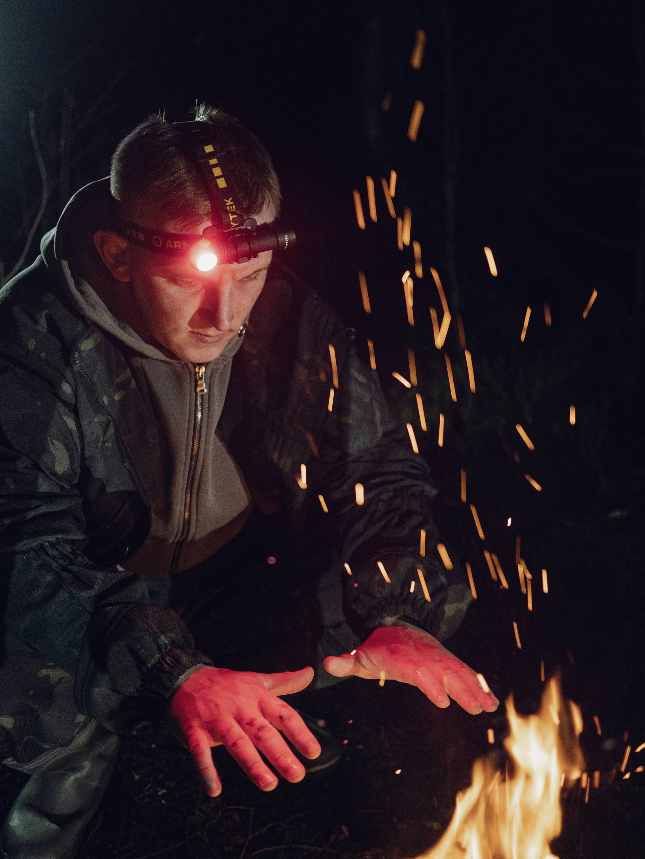Mann mit Stirnlampe wärmt sich nachts an einem Lagerfeuer, Funken fliegen in die Luft. Er trägt dunkle Kleidung und schaut konzentriert.