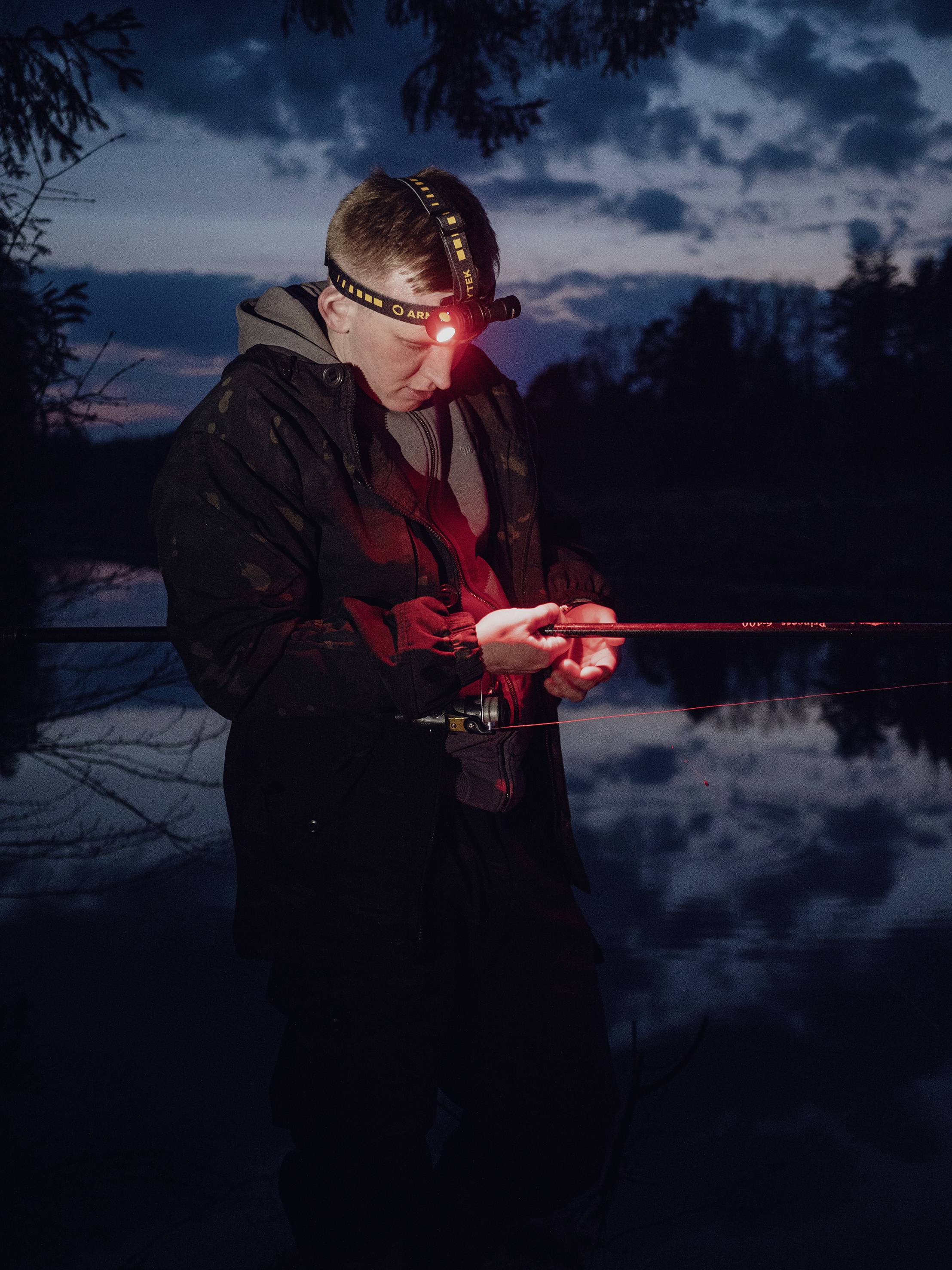 Eine Person steht am Wasser und überprüft eine Angelrute, beleuchtet von einem Stirnlicht in der Abenddämmerung.