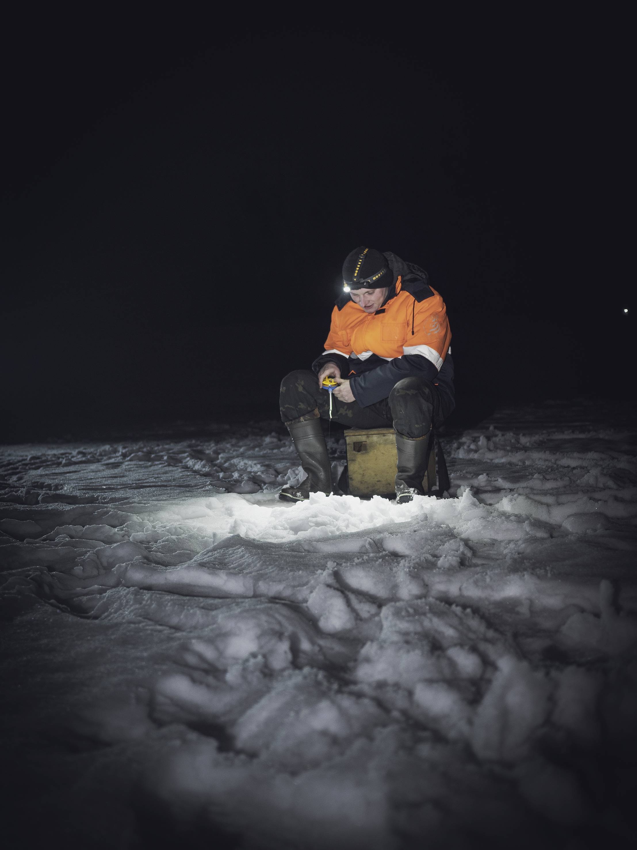 Eine Person sitzt nachts auf einem Hocker im Schnee, trägt eine orange Jacke und eine Stirnlampe, konzentriert sich auf ihre Hände.
