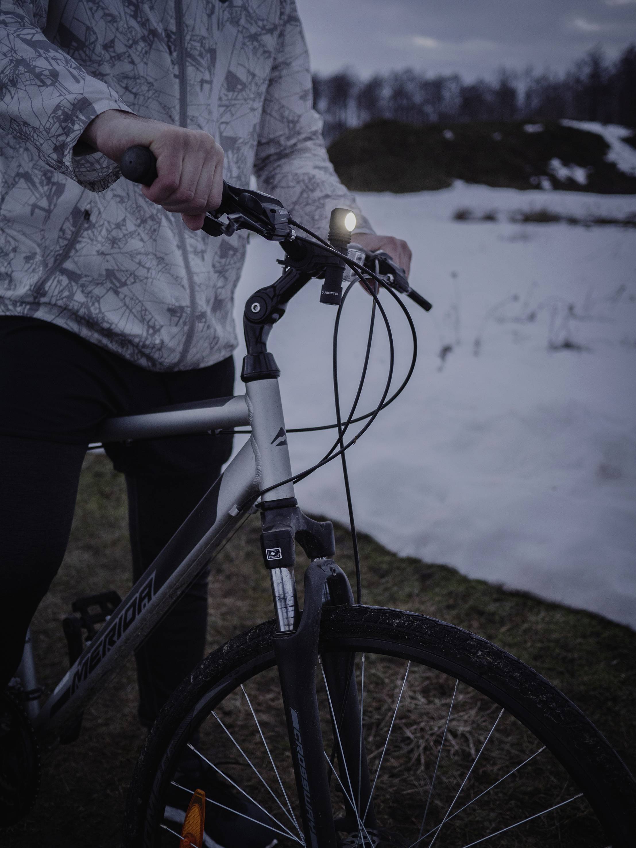 Eine Person steht im Freien neben einem Fahrrad. Im Hintergrund ist verschneite Landschaft zu sehen, es scheint dämmerig zu sein.