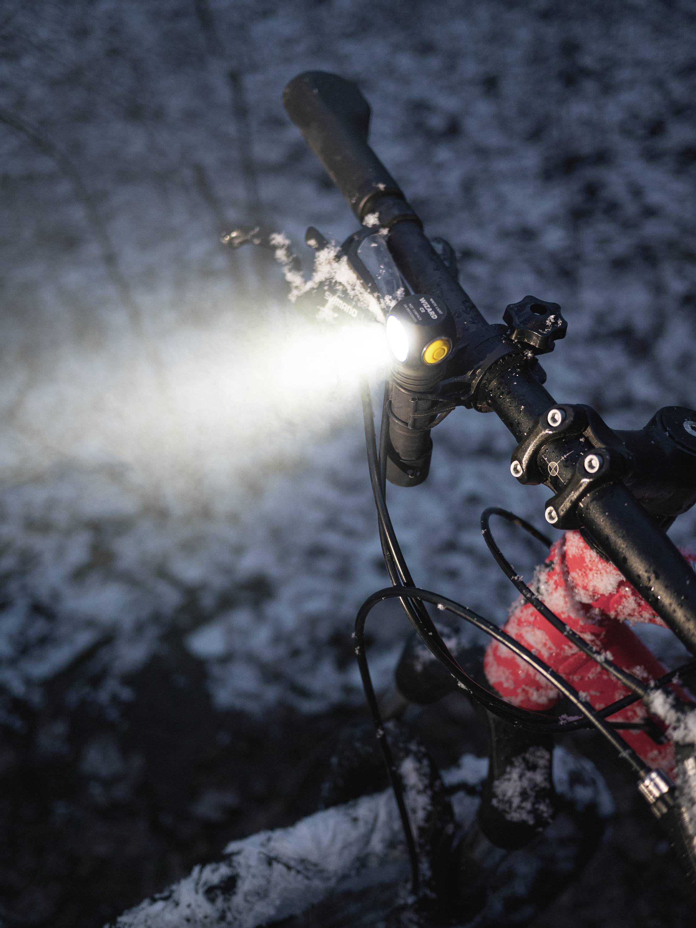 Fahrradlenker im Schnee mit eingeschaltetem Frontlicht, das den Weg beleuchtet. Winterliche Umgebung, Lichter reflektieren.