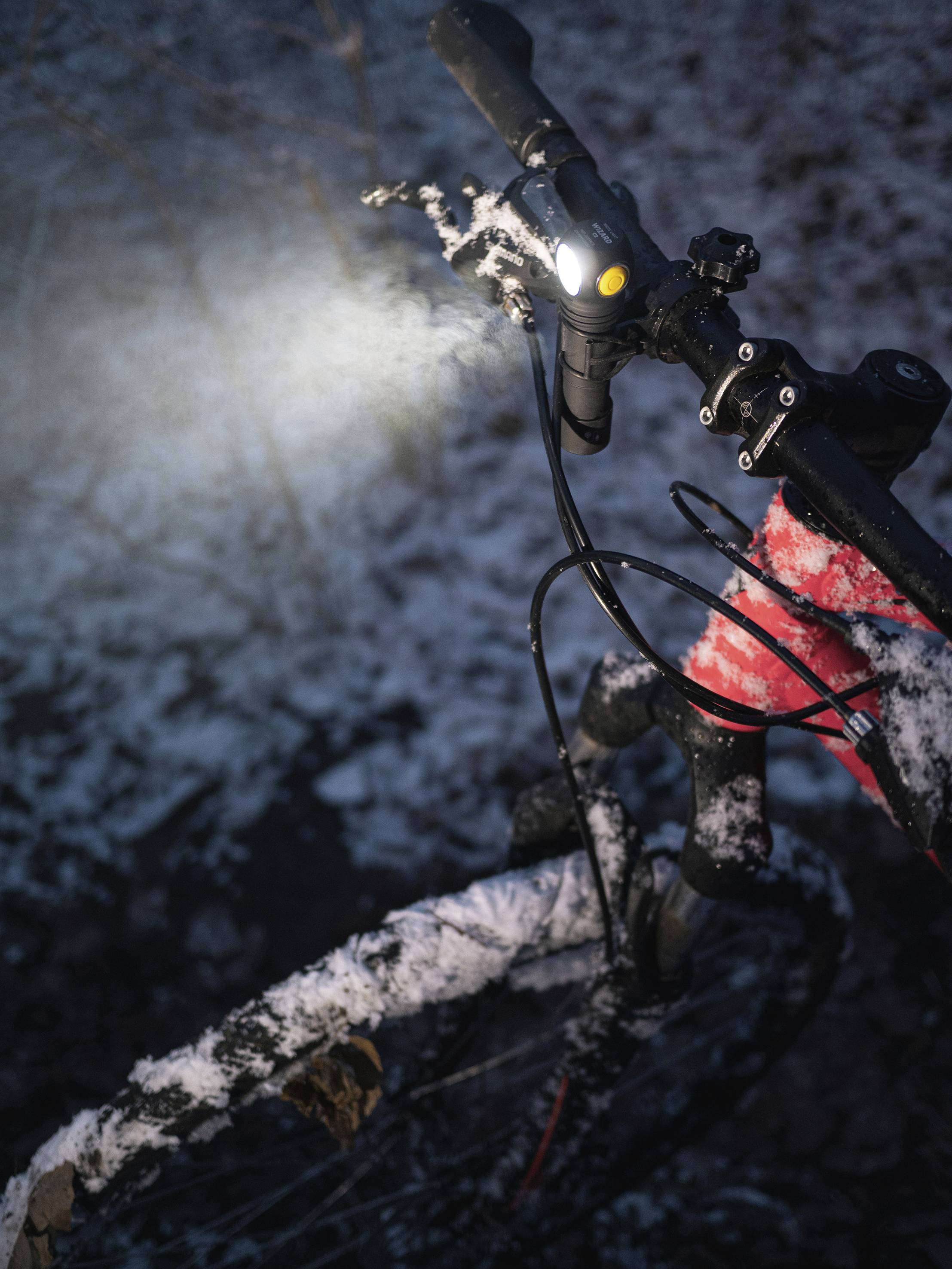 Ein Fahrrad mit eingeschaltetem Frontlicht steht im Schnee. Der Rahmen ist rot, der Lenker ist leicht mit Schnee bedeckt. Die Umgebung ist dunkel.