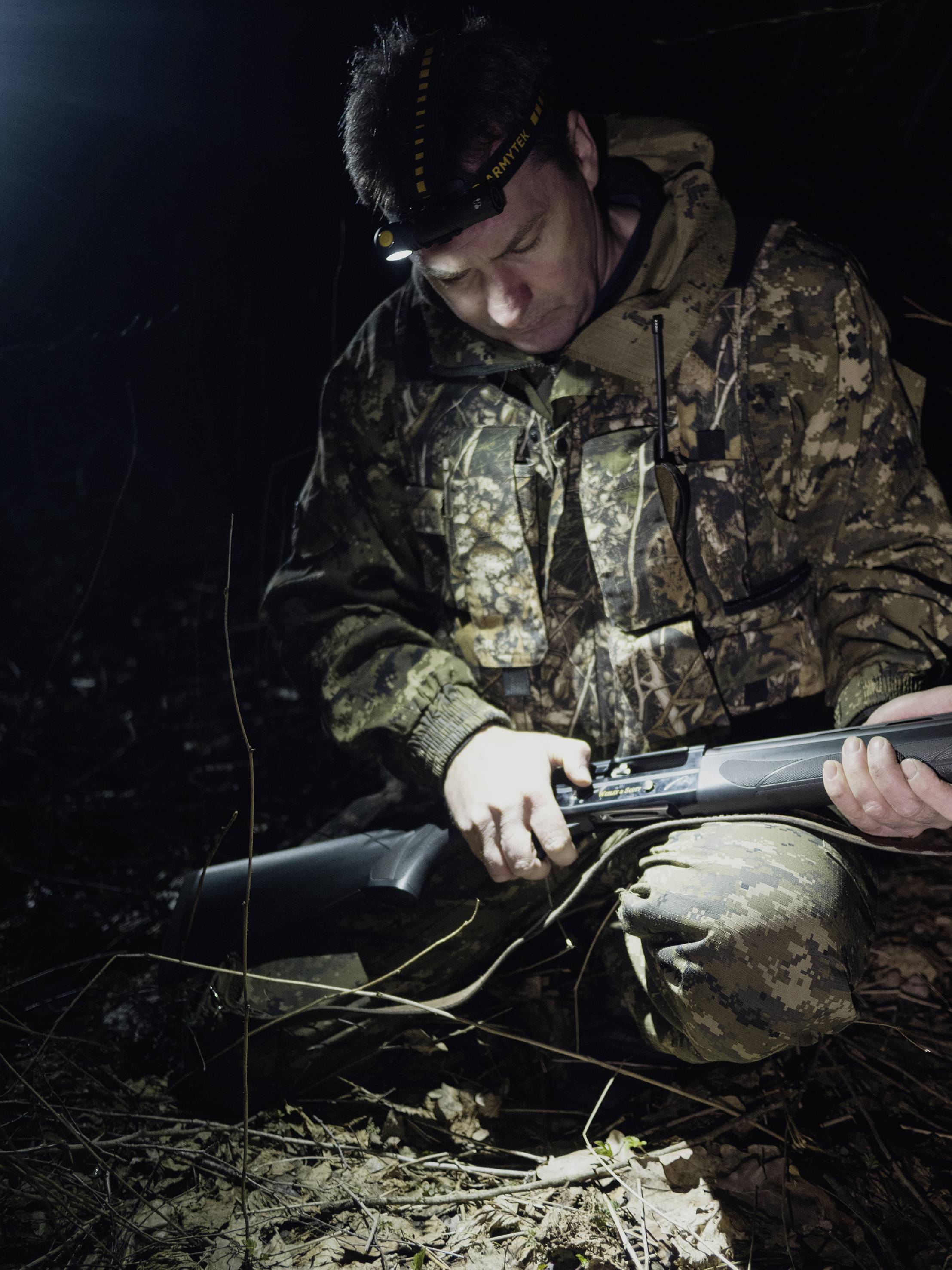 Eine Person in Tarnkleidung kniet im Dunkeln und überprüft eine Waffe mit einer Stirnlampe. Die Szene wirkt angespannt und fokussiert.
