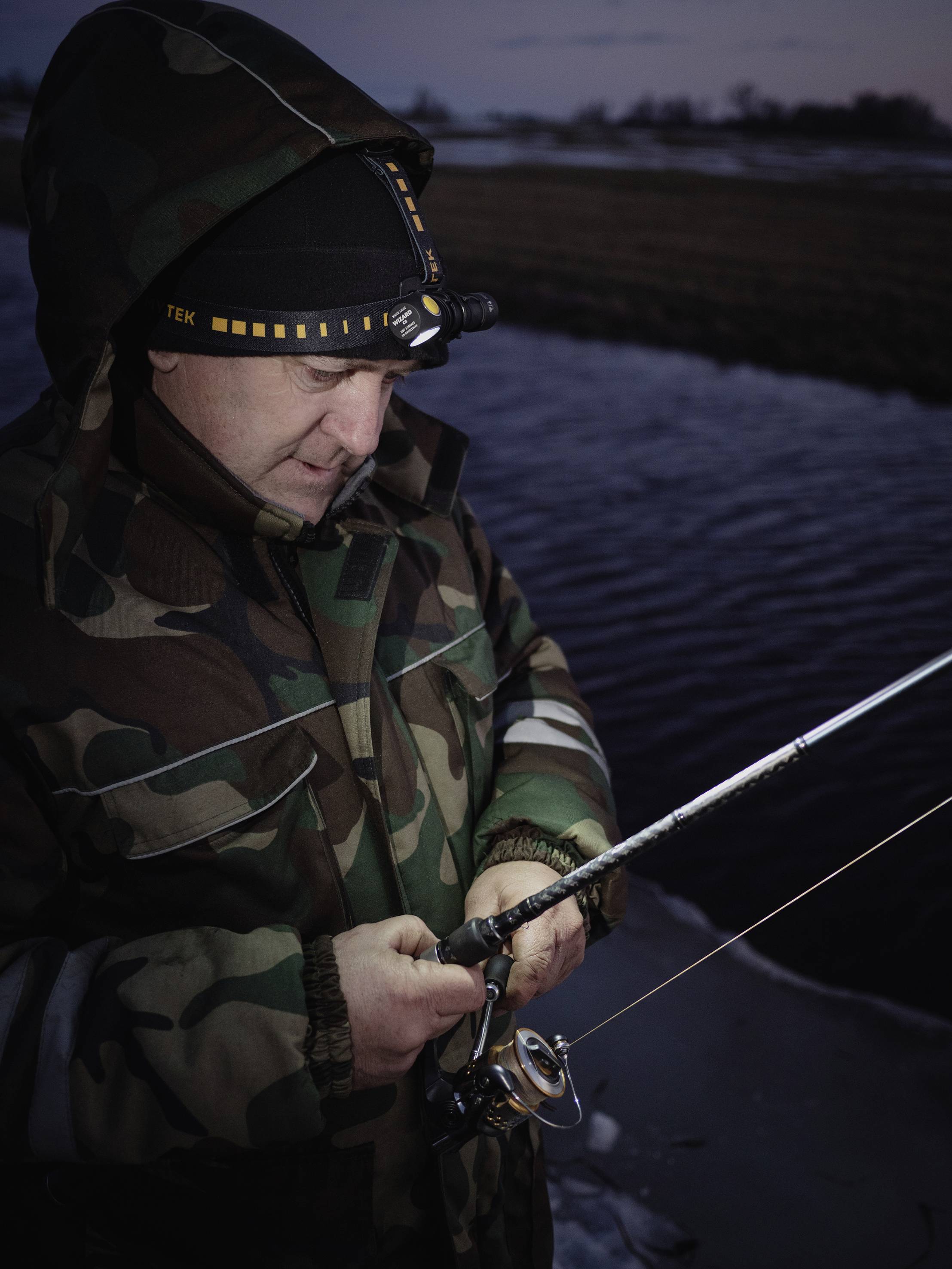 Ein Angler in Tarnkleidung steht am Ufer eines Gewässers in der Dämmerung, konzentriert auf das Einstellen seiner Angelrute.
