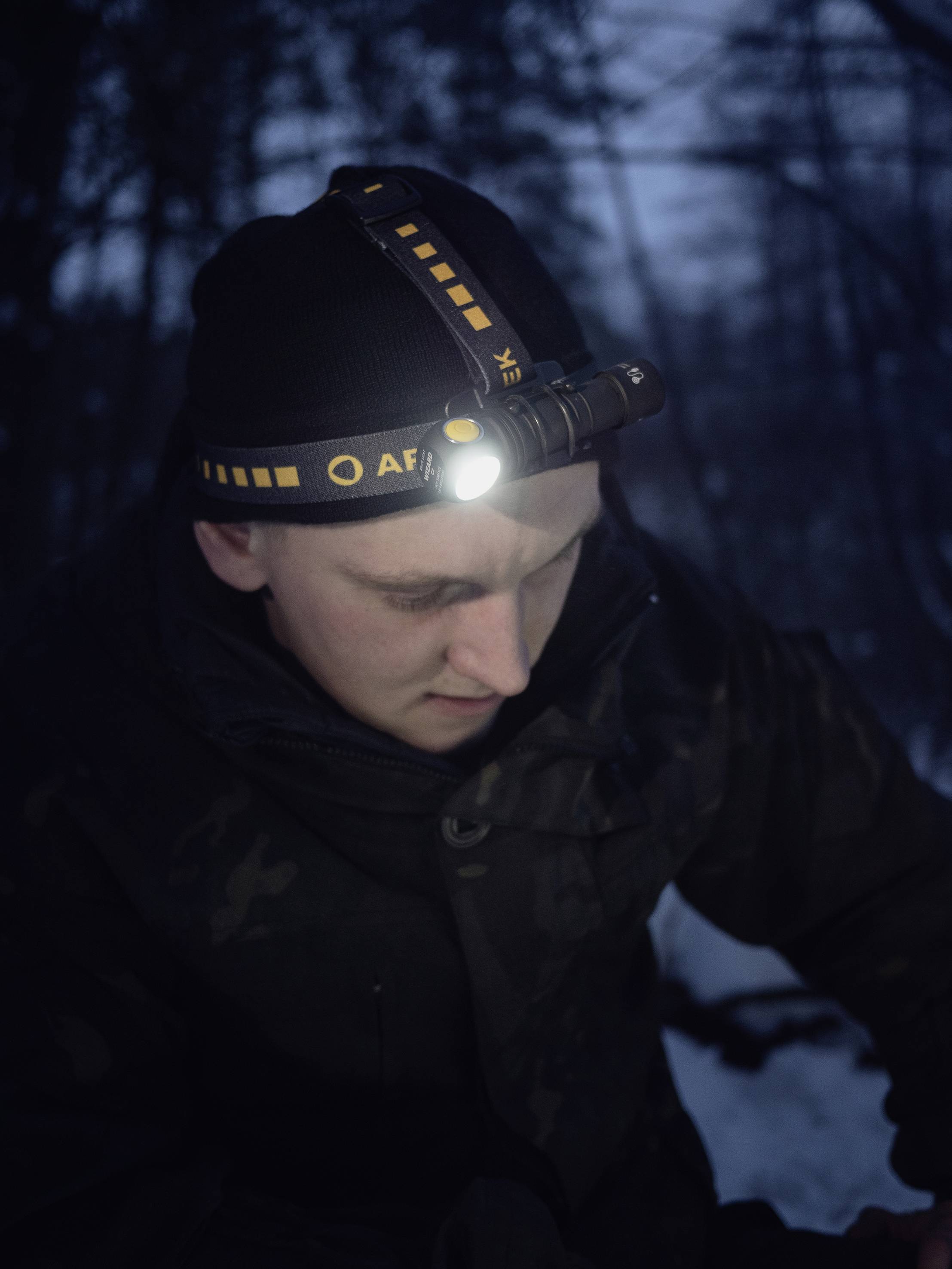 Eine Person im Wald bei Nacht, trägt eine Stirnlampe, die ihr Gesicht beleuchtet. Sie ist warm angezogen und konzentriert sich auf etwas.