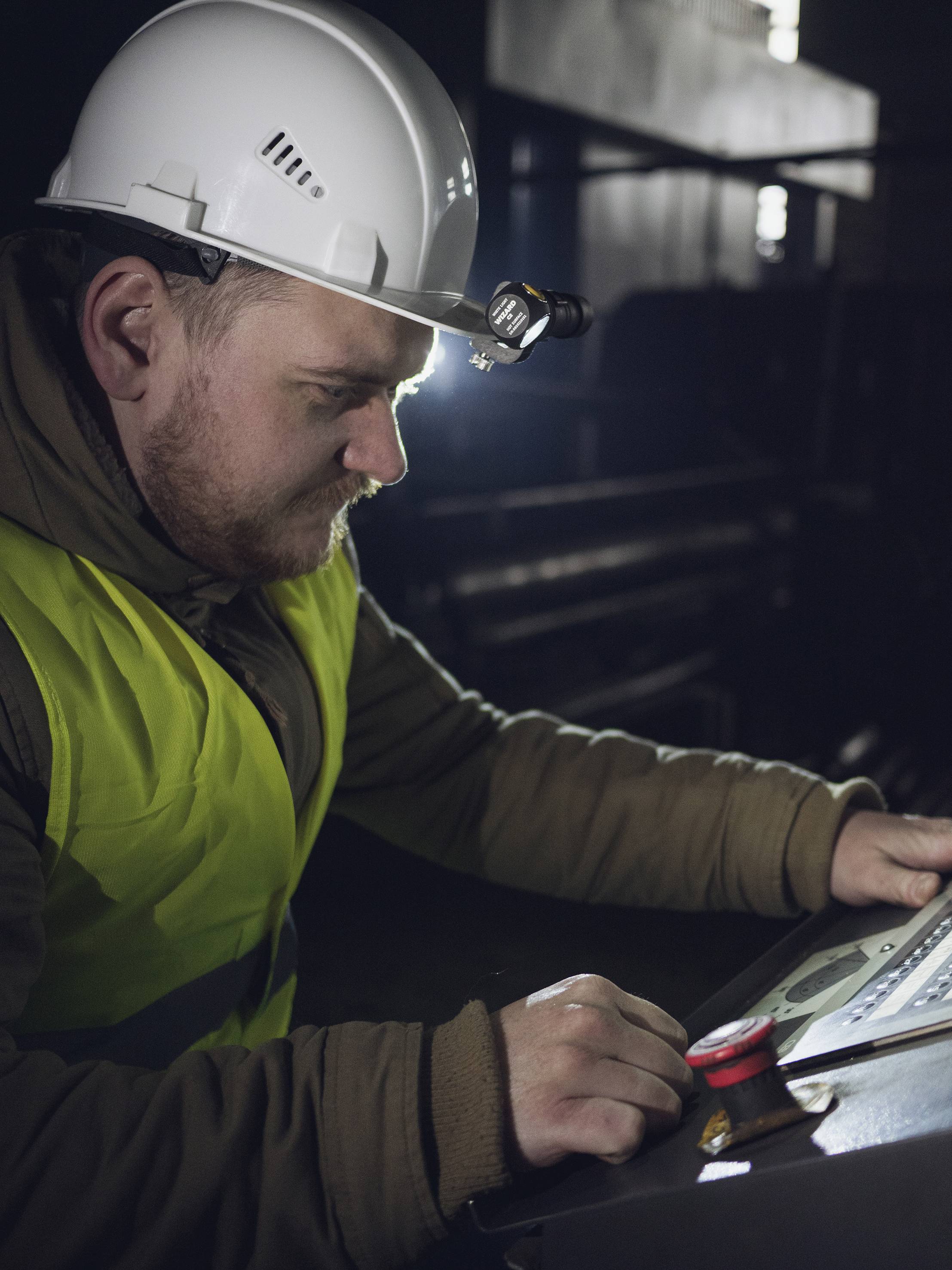 Ein Arbeiter mit Helm und Warnweste bedient im Halbdunkel eine Maschine, fokussiert auf ein Bedienfeld mit Touchscreen.