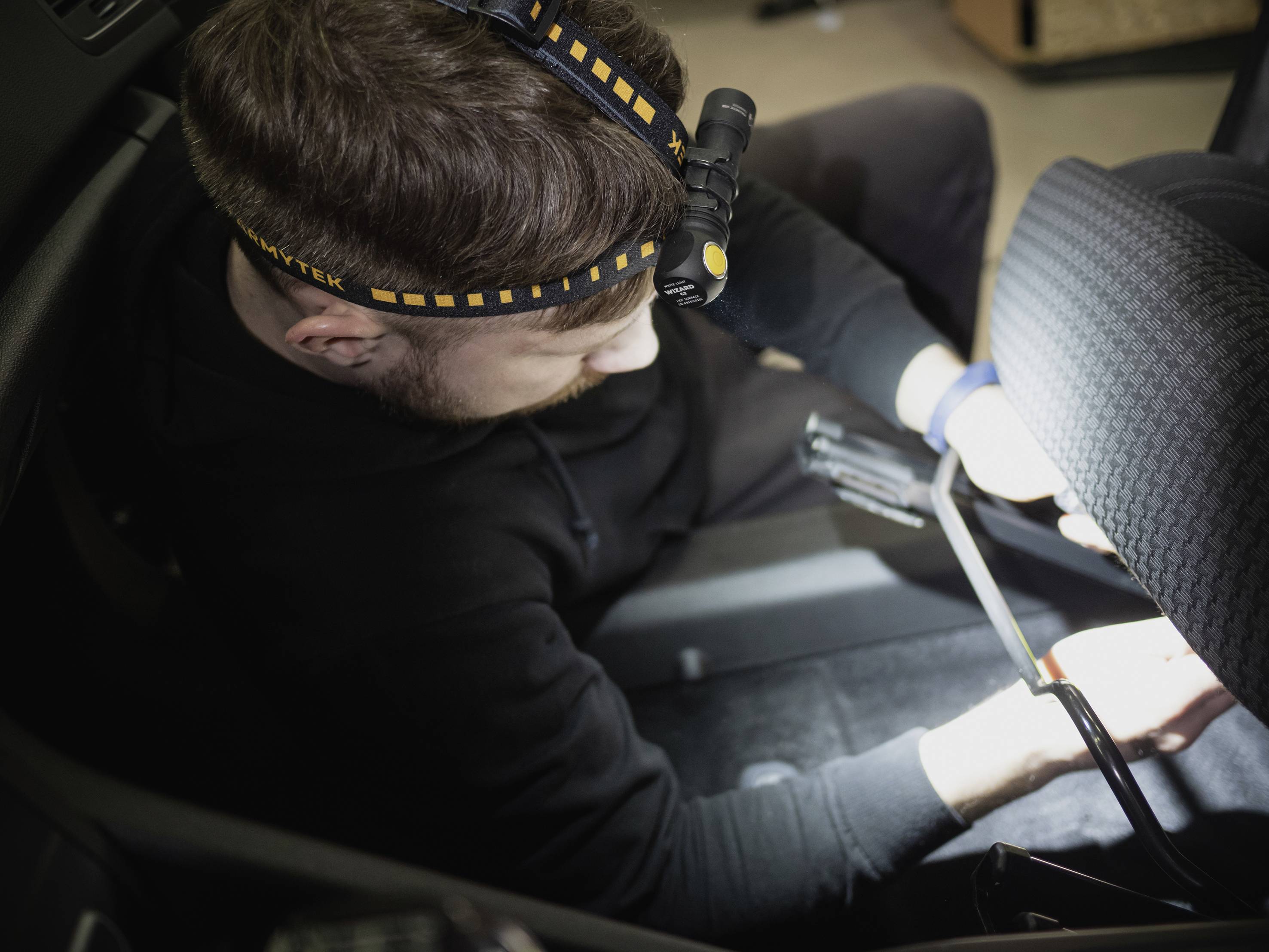 Ein Mann im schwarzen Pullover arbeitet mit einem grellen Stirnlampe im Inneren eines Autos, konzentriert auf die Reparatur eines Sitzes.