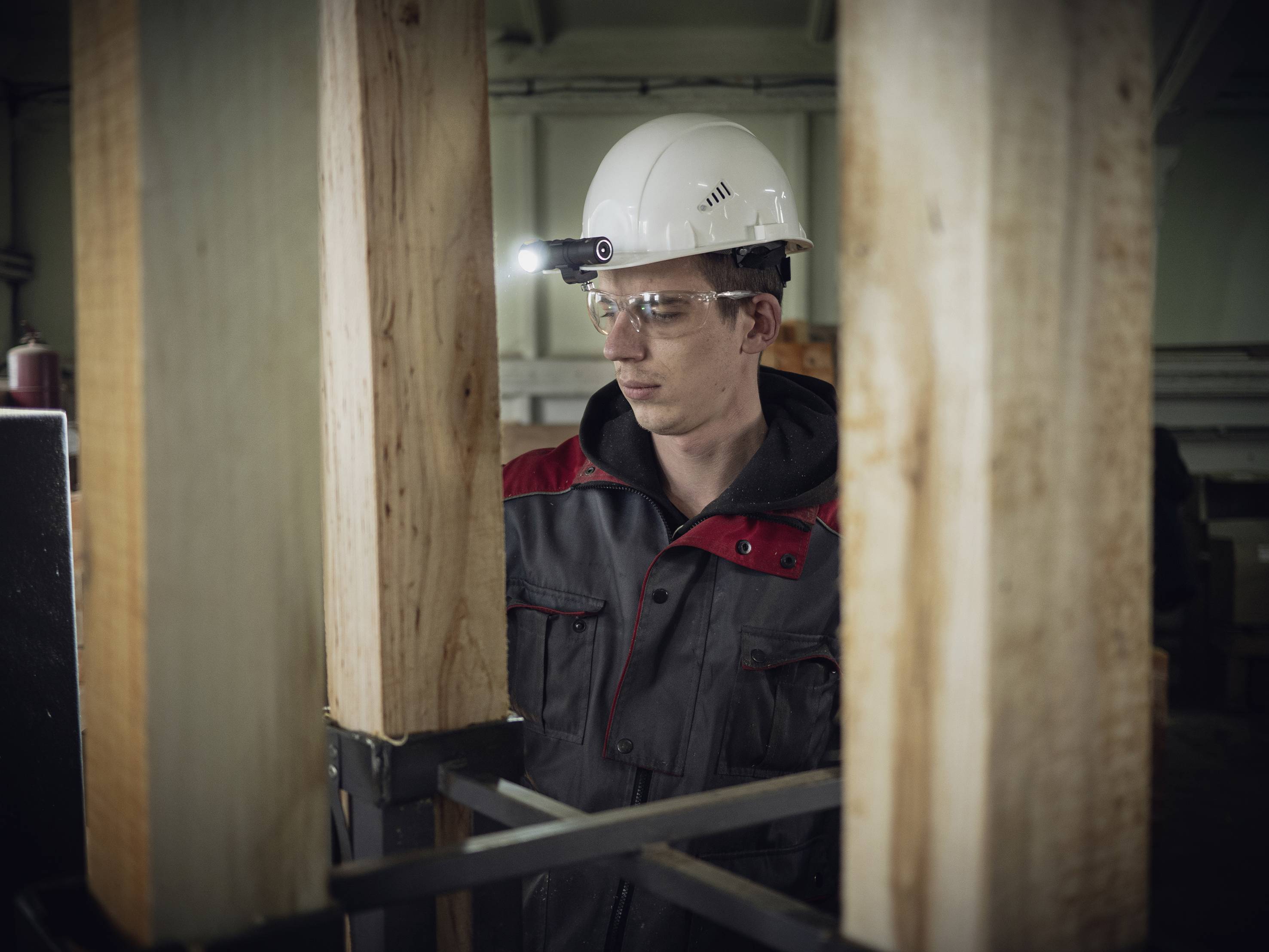 Ein Mann mit Schutzbrille und Helm untersucht Holzpfähle in einer Werkstatt, beleuchtet von einer Stirnlampe.