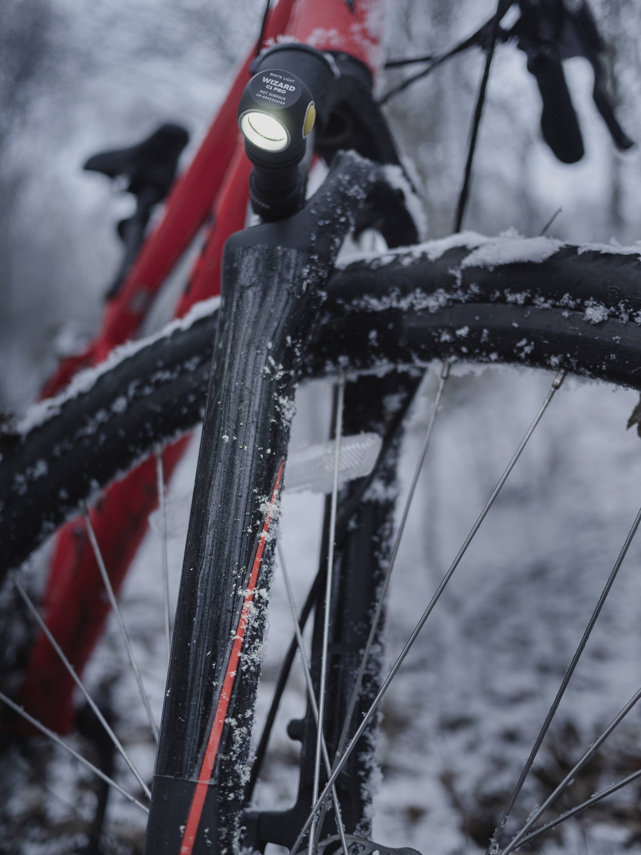 Nahaufnahme eines roten Fahrrads im Schnee, fokussiert auf das Vorderrad und das eingeschaltete Vorderlicht.