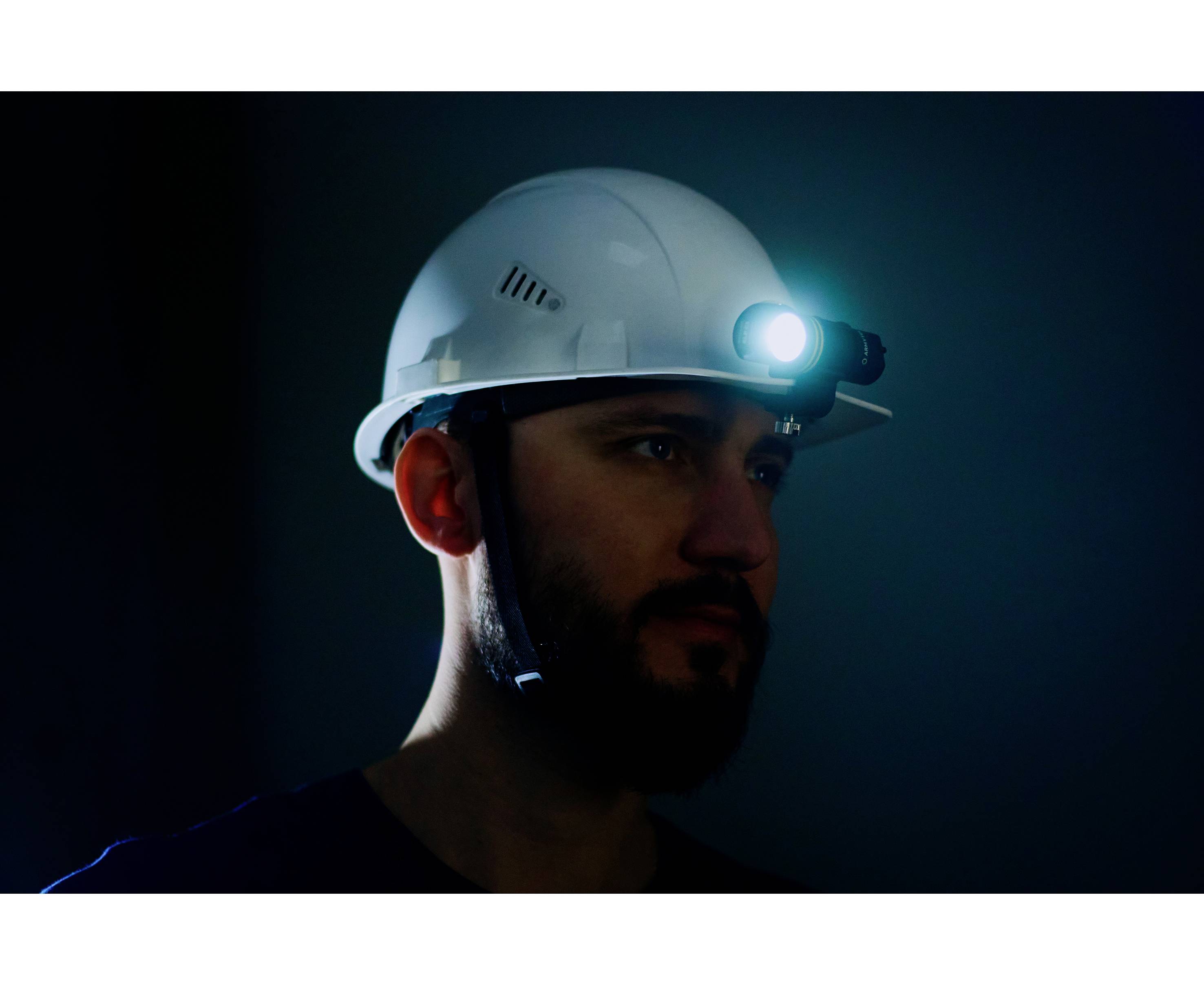 Ein Mann mit Helm und eingeschalteter Stirnlampe steht in dunkler Umgebung, beleuchtet sein Gesicht leicht seitlich.