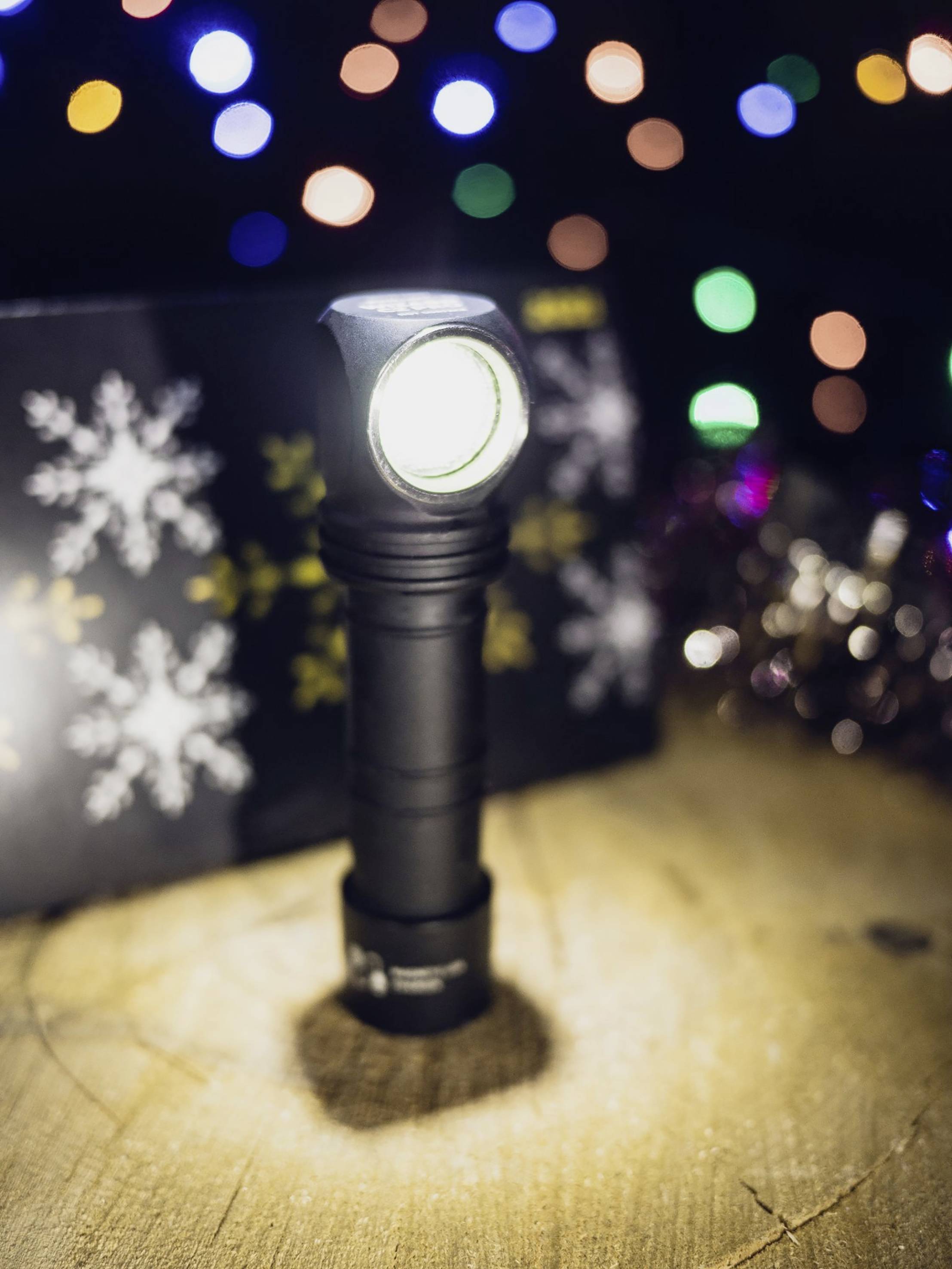 Eine schwarze Taschenlampe steht auf einem Holztisch mit bunten Lichtern im Hintergrund. Sie strahlt helles Licht aus.