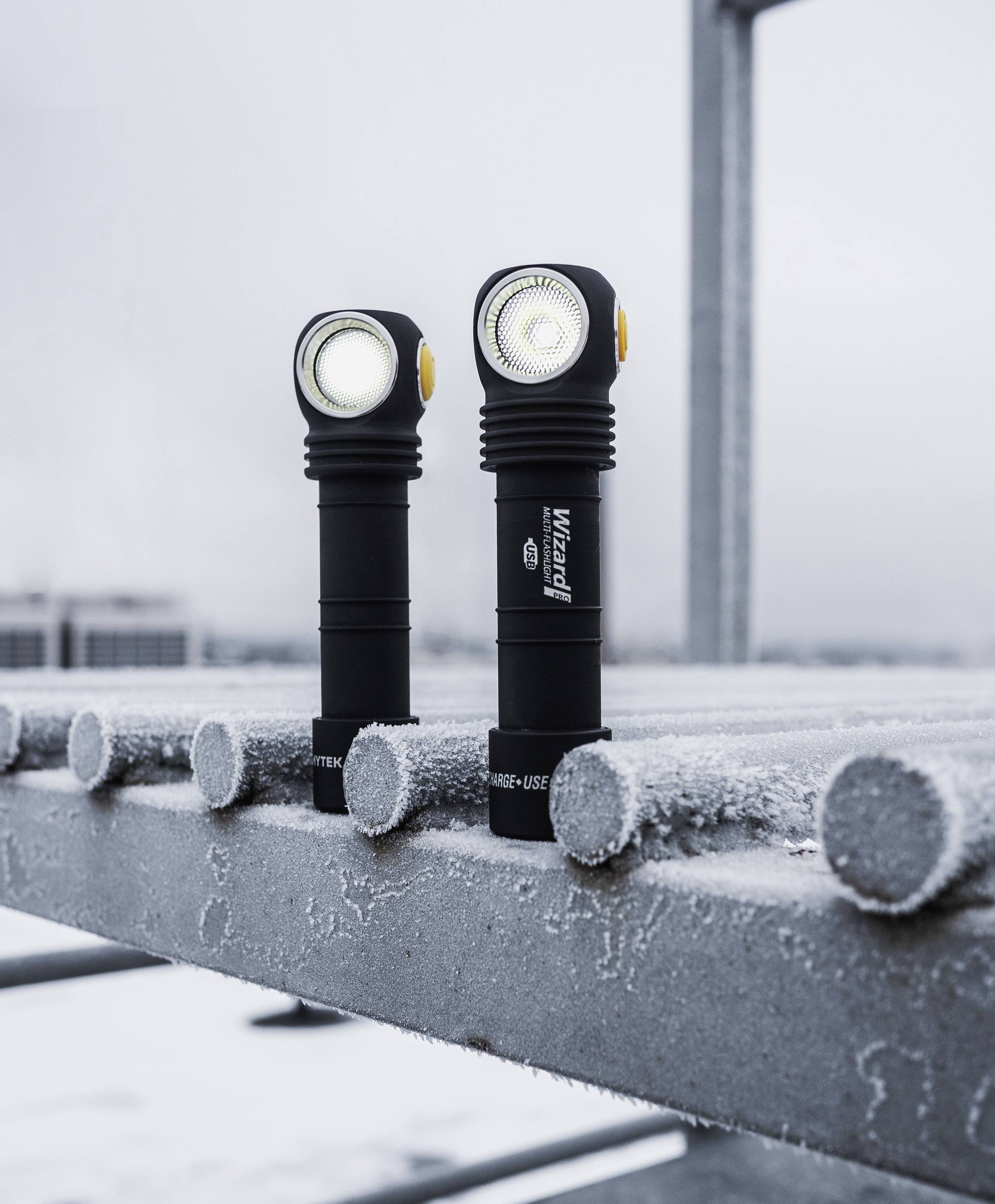 Zwei taktische Taschenlampen stehen auf einem vereisten Metallgitter, leuchten in unterschiedliche Richtungen. Hintergrund: verschwommene Gebäude.