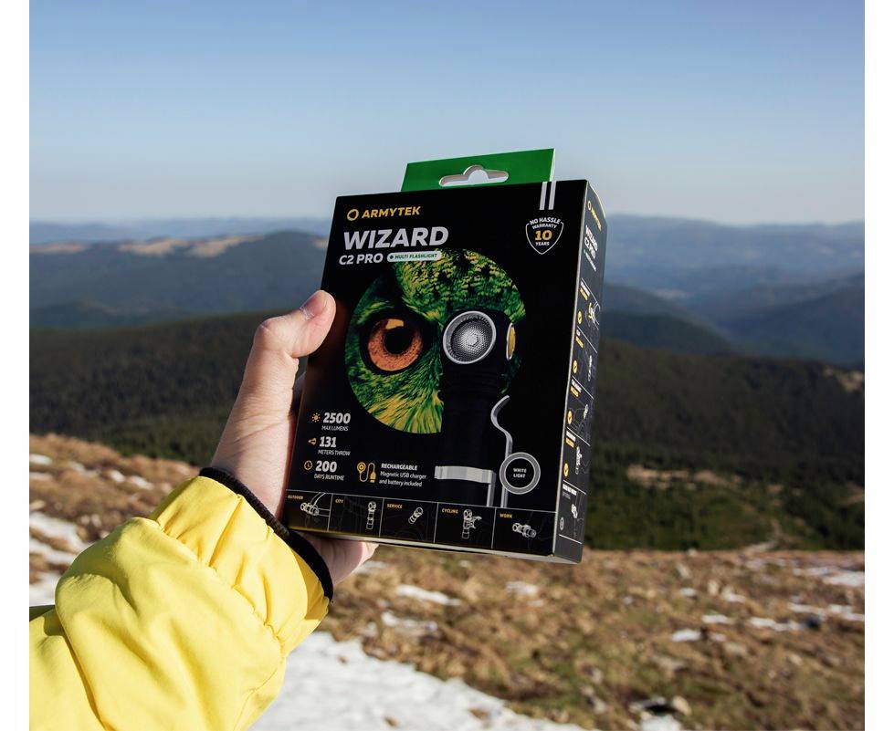 Eine Person hält eine 'Armytek Wizard C2 Pro' Taschenlampe-Schachtel vor einer Berglandschaft in der Hand.