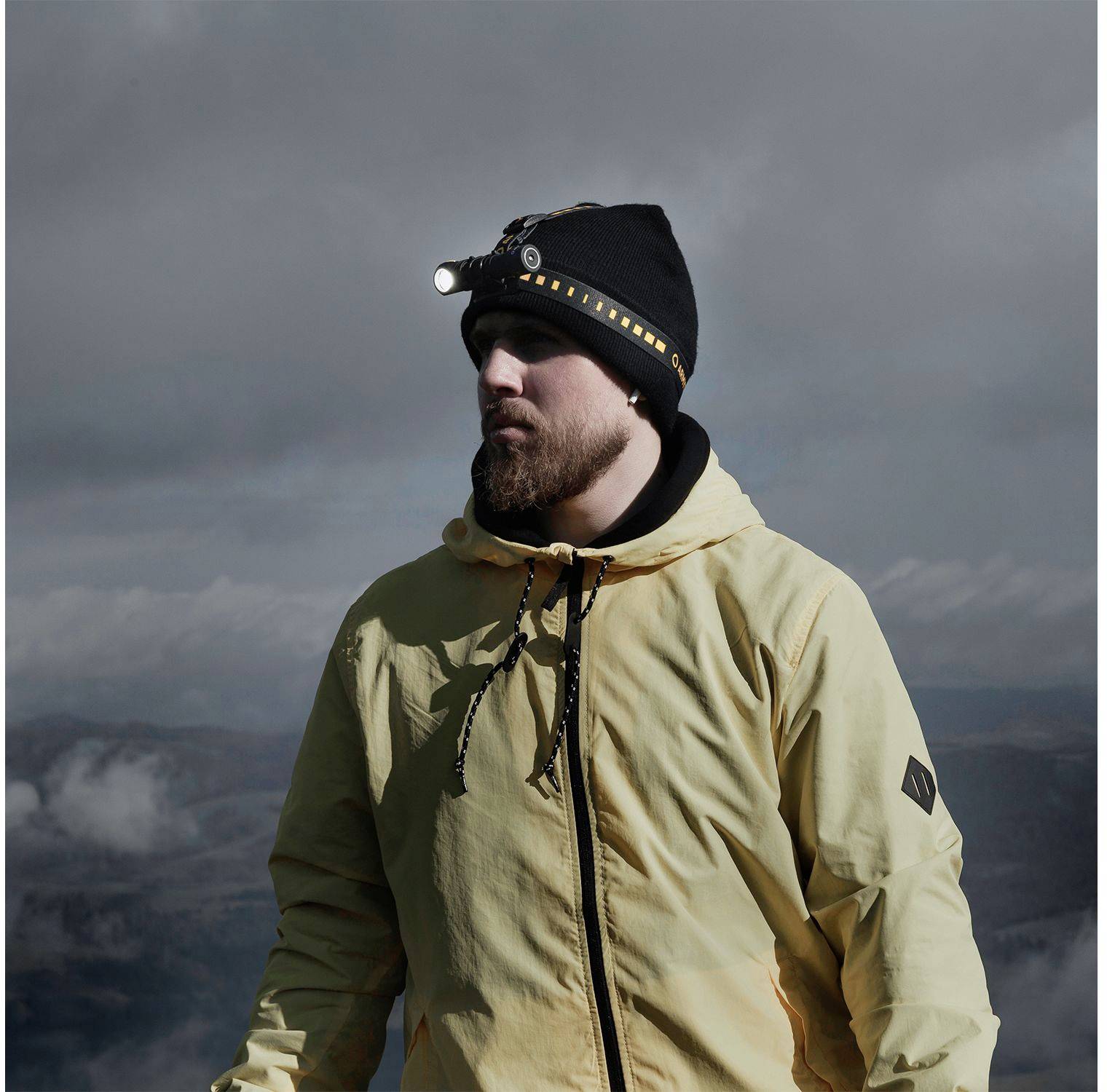 Eine Person in Outdoor-Kleidung mit Stirnlampe steht vor einem wolkigen Himmel und sieht entschlossen aus, als ob sie wandert oder erkundet.