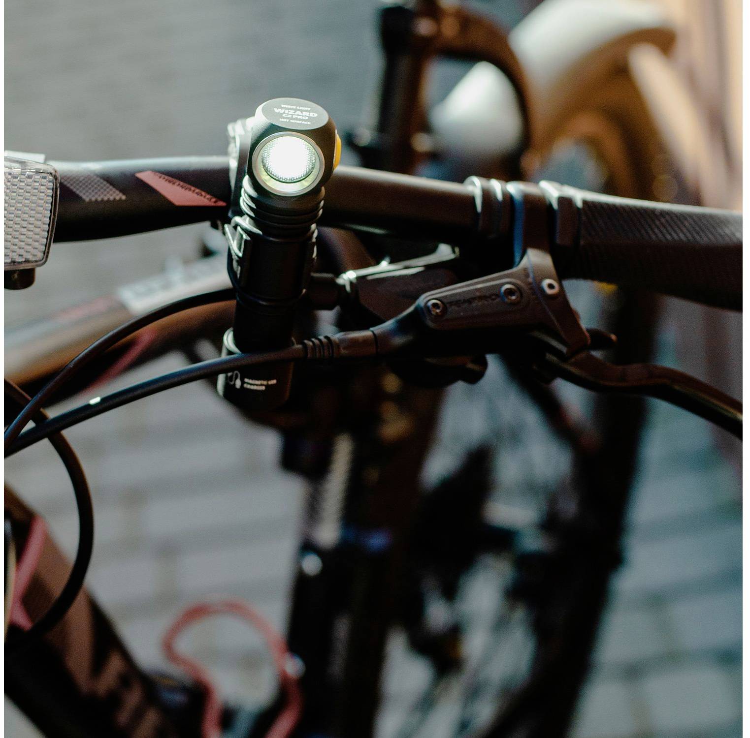 Fahrradlenker mit eingeschaltetem Licht in der Dämmerung, detaillierte Ansicht der Bremsen und Kabel, im Hintergrund leicht unscharf.