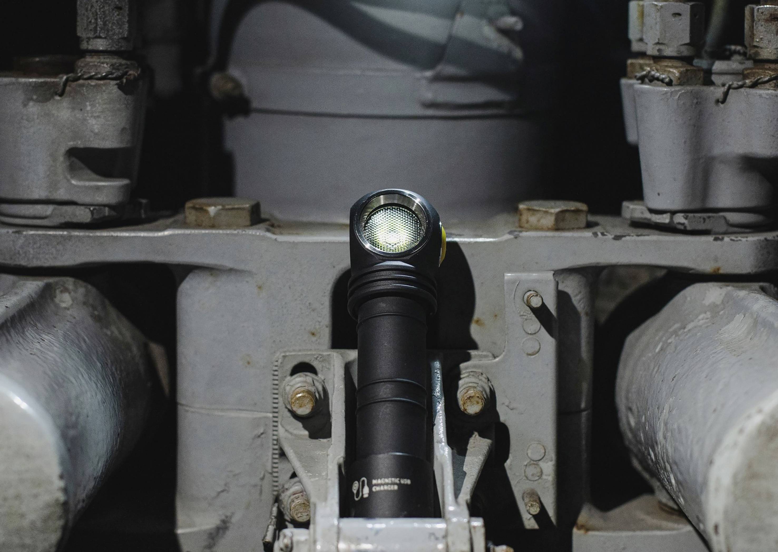 Taschenlampe liegt auf Metallstruktur unter Rohren, leuchtet nach vorne. Umgebung wirkt industriell und robust.