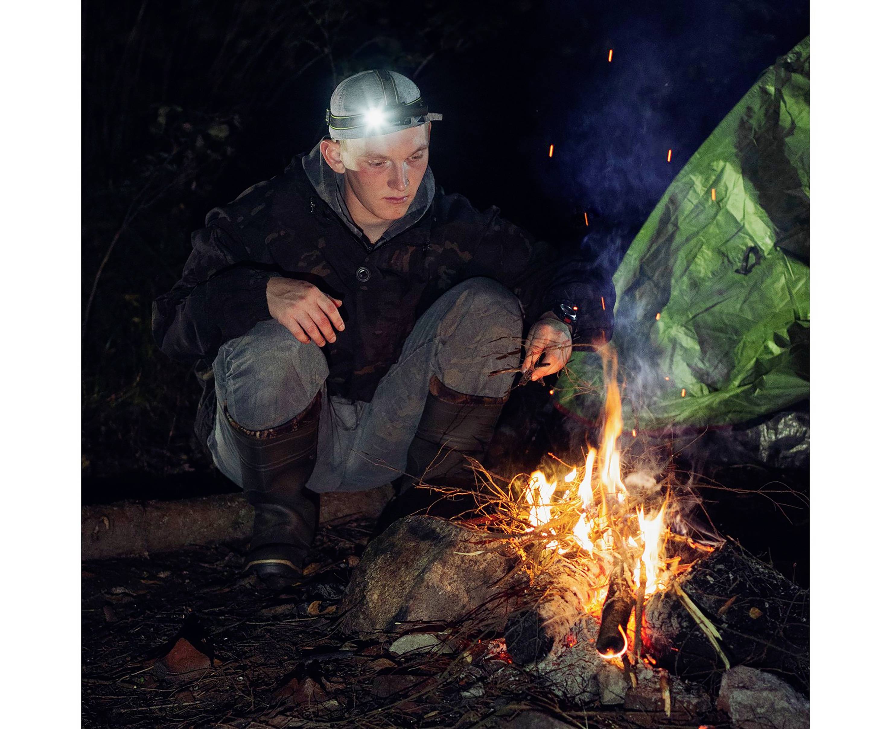 Eine Person mit Stirnlampe hockt nachts am Lagerfeuer und beobachtet die Flammen. Im Hintergrund ist eine grüne Plane zu sehen.