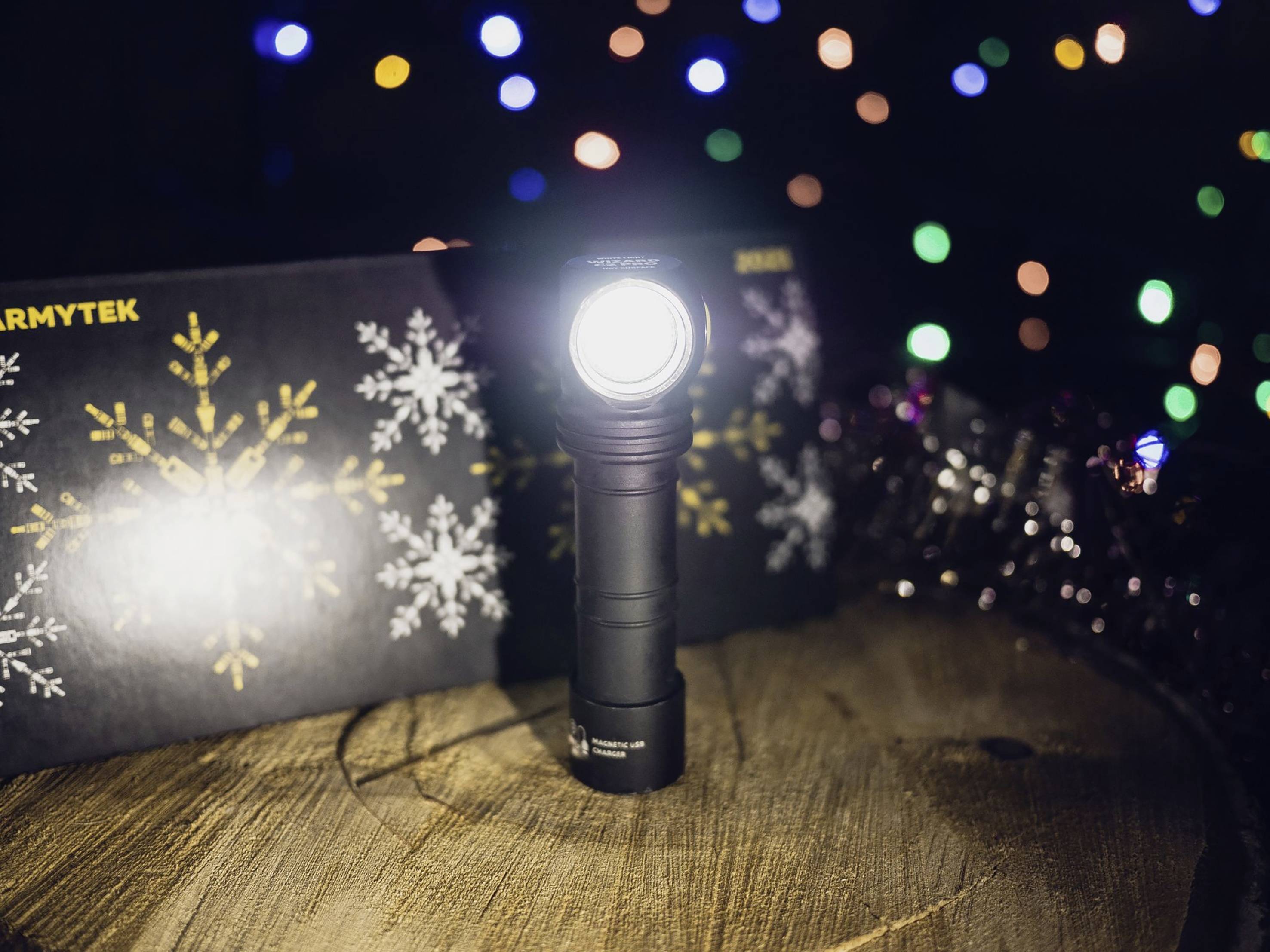 Eine Taschenlampe leuchtet hell auf einem Holztisch. Im Hintergrund sind unscharfe, bunte Lichter und eine schwarze Schachtel mit Schneeflocken.