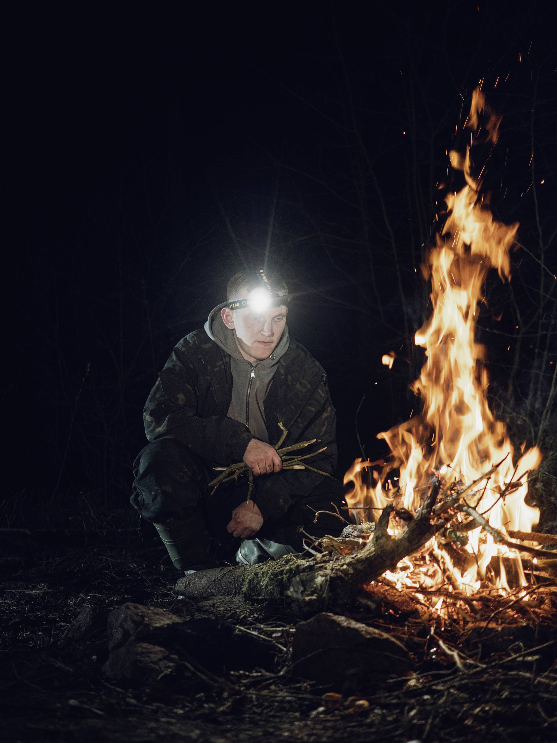 Person mit Stirnlampe kniet in der Nacht neben einem großen Lagerfeuer im Wald. Er legt Zweige ins Feuer.