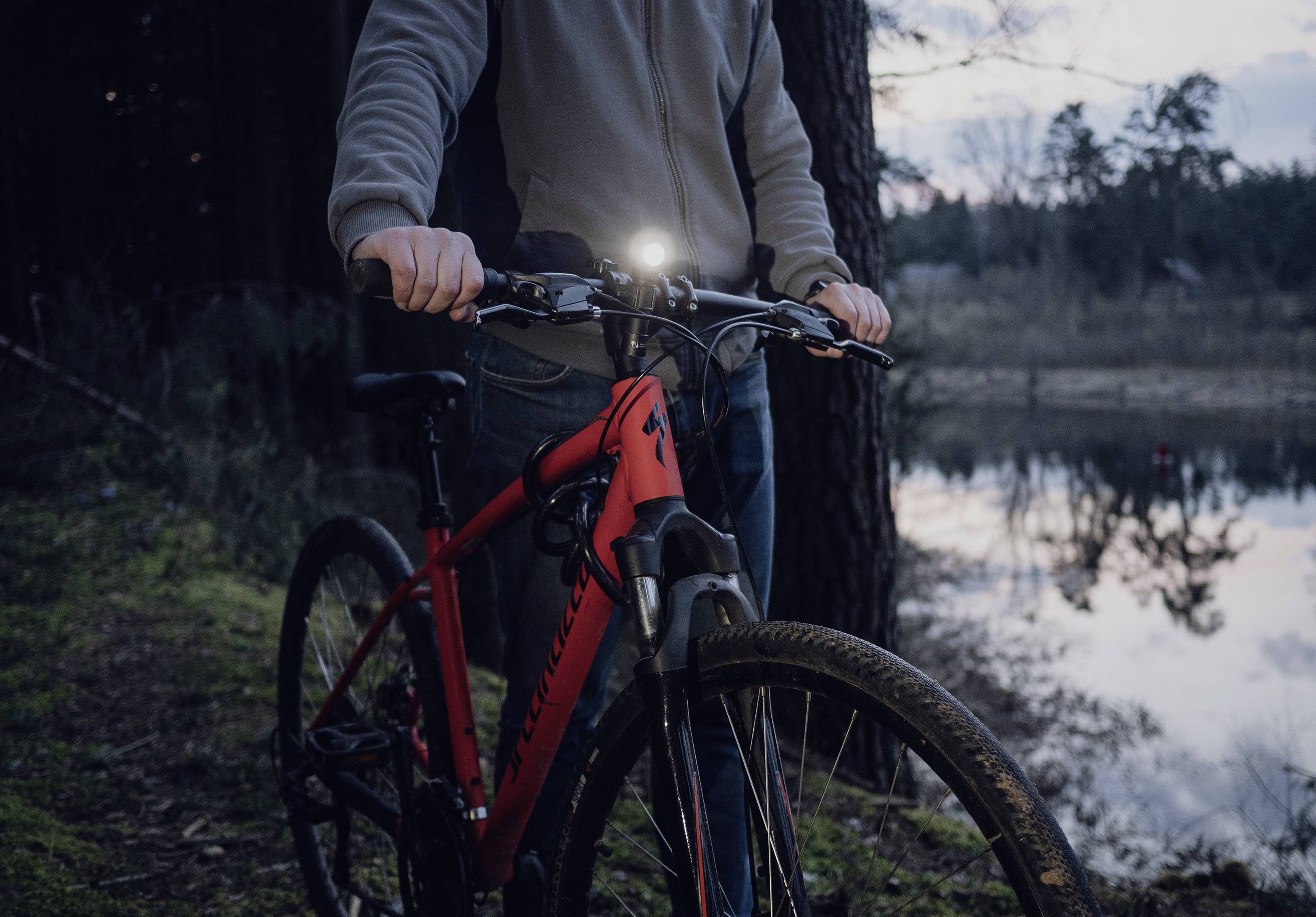 Eine Person hält ein rotes Fahrrad mit eingeschaltetem Vorderlicht. Im Hintergrund ist ein Wald mit einem Teich bei Dämmerung zu sehen.
