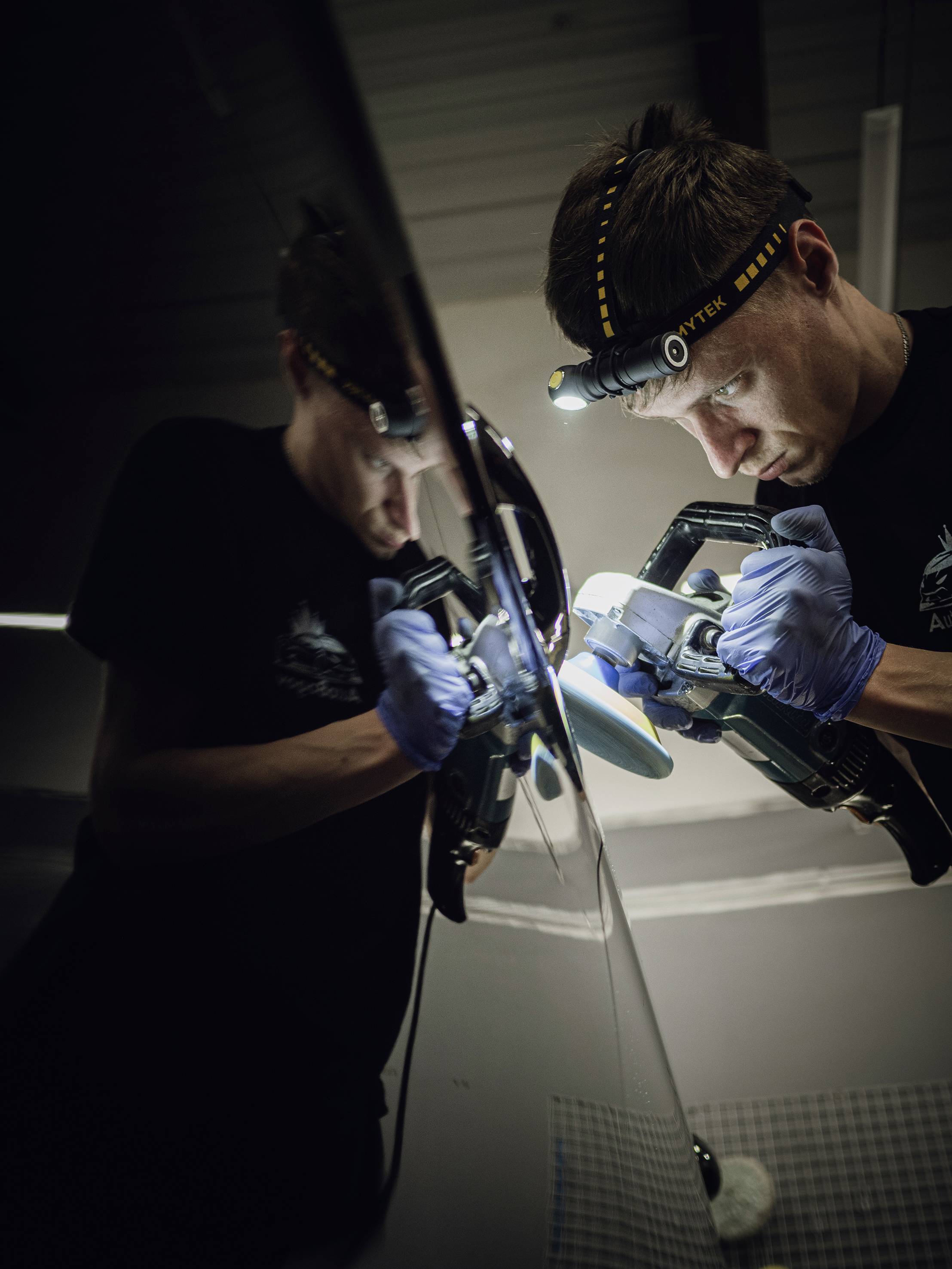 Ein Mann mit Stirnlampe und Handschuhen poliert ein Autoteil, das sich spiegelt. Er arbeitet konzentriert und präzise.