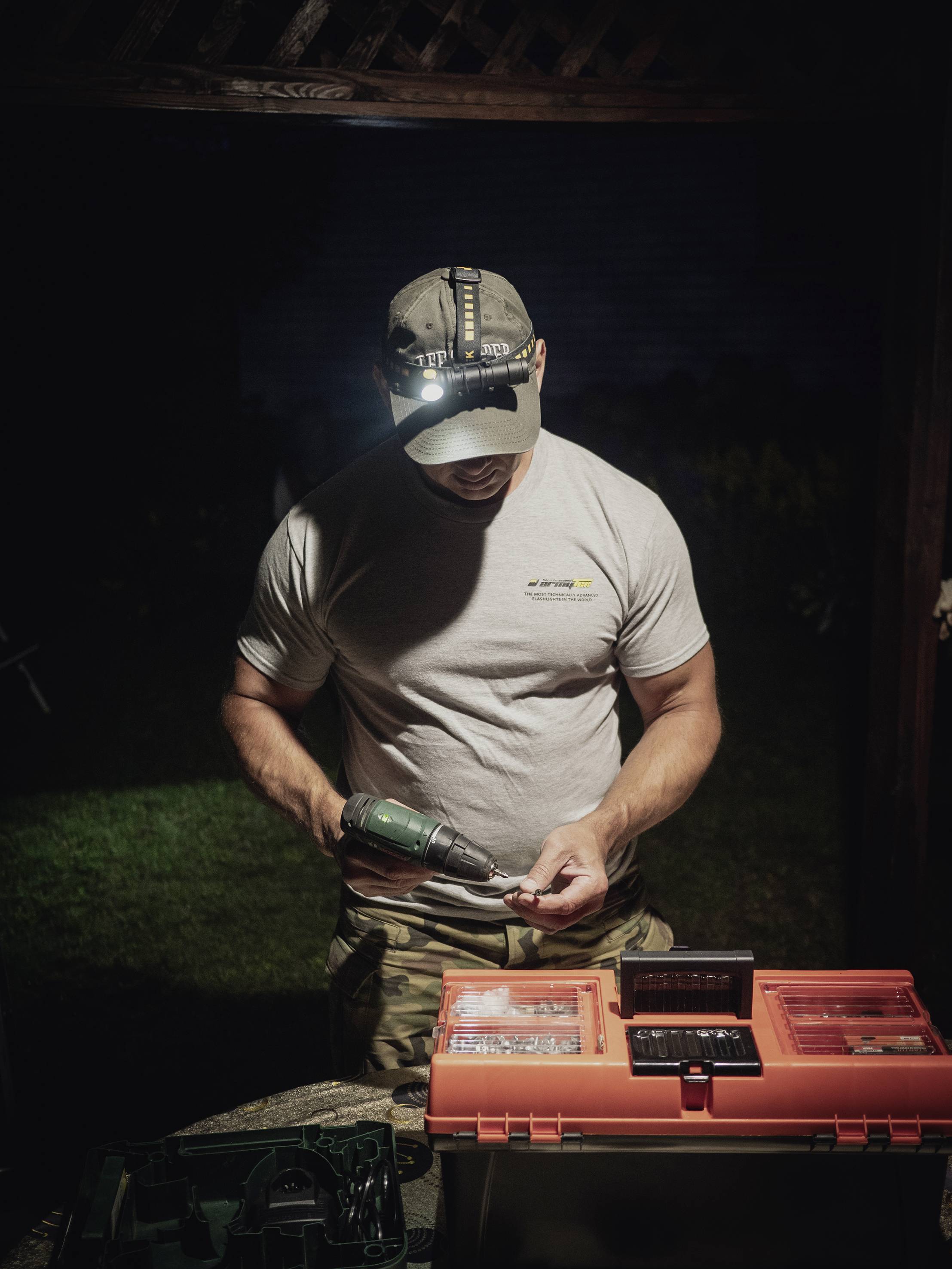 Ein Mann arbeitet im Freien bei Nacht mit einer Bohrmaschine neben einem geöffneten Werkzeugkasten. Er trägt ein T-Shirt und eine Mütze.