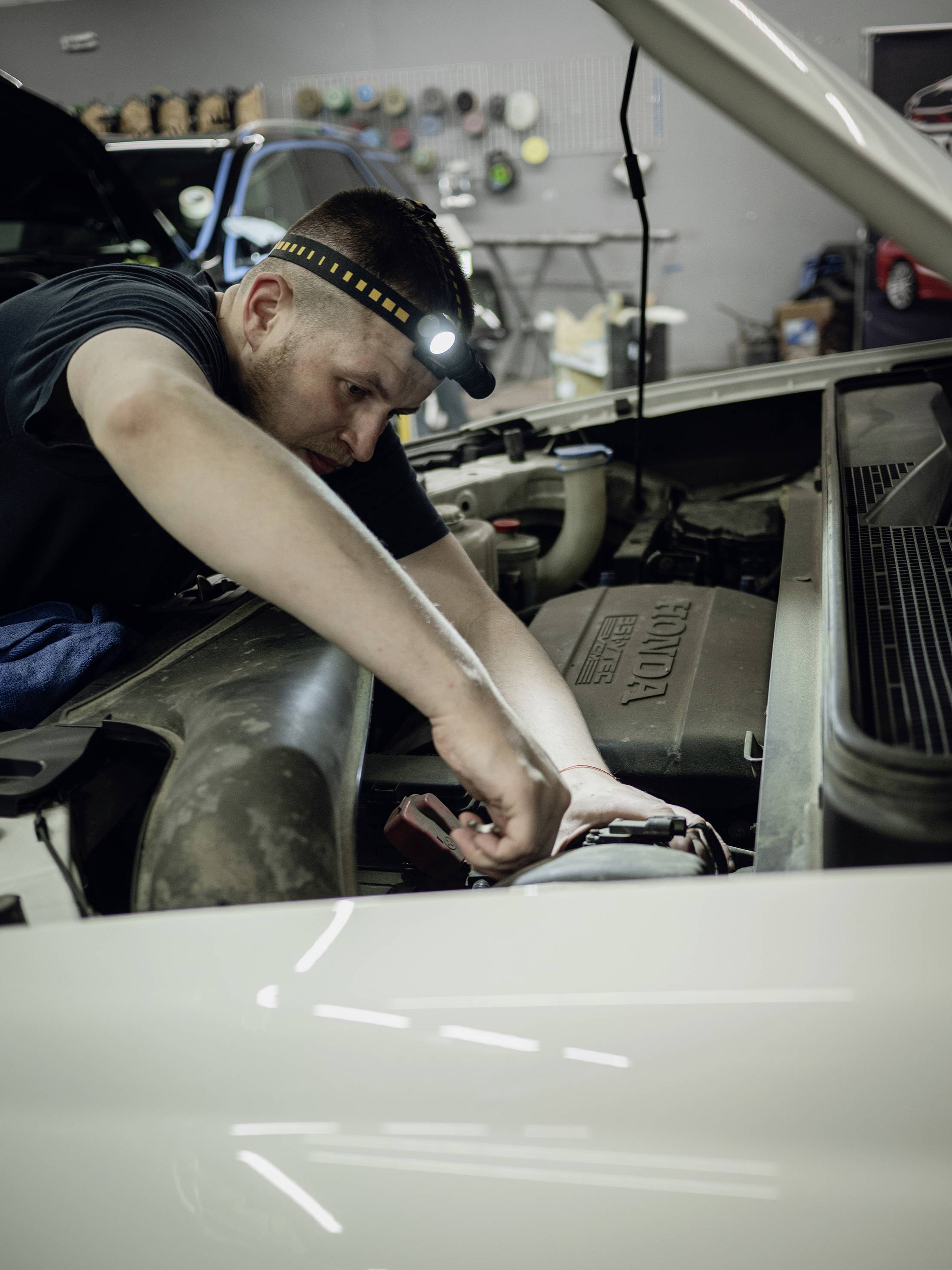 Eine Person arbeitet unter der geöffneten Motorhaube eines Autos, konzentriert, mit einer Stirnlampe ausgestattet, am Fahrzeugmotor.