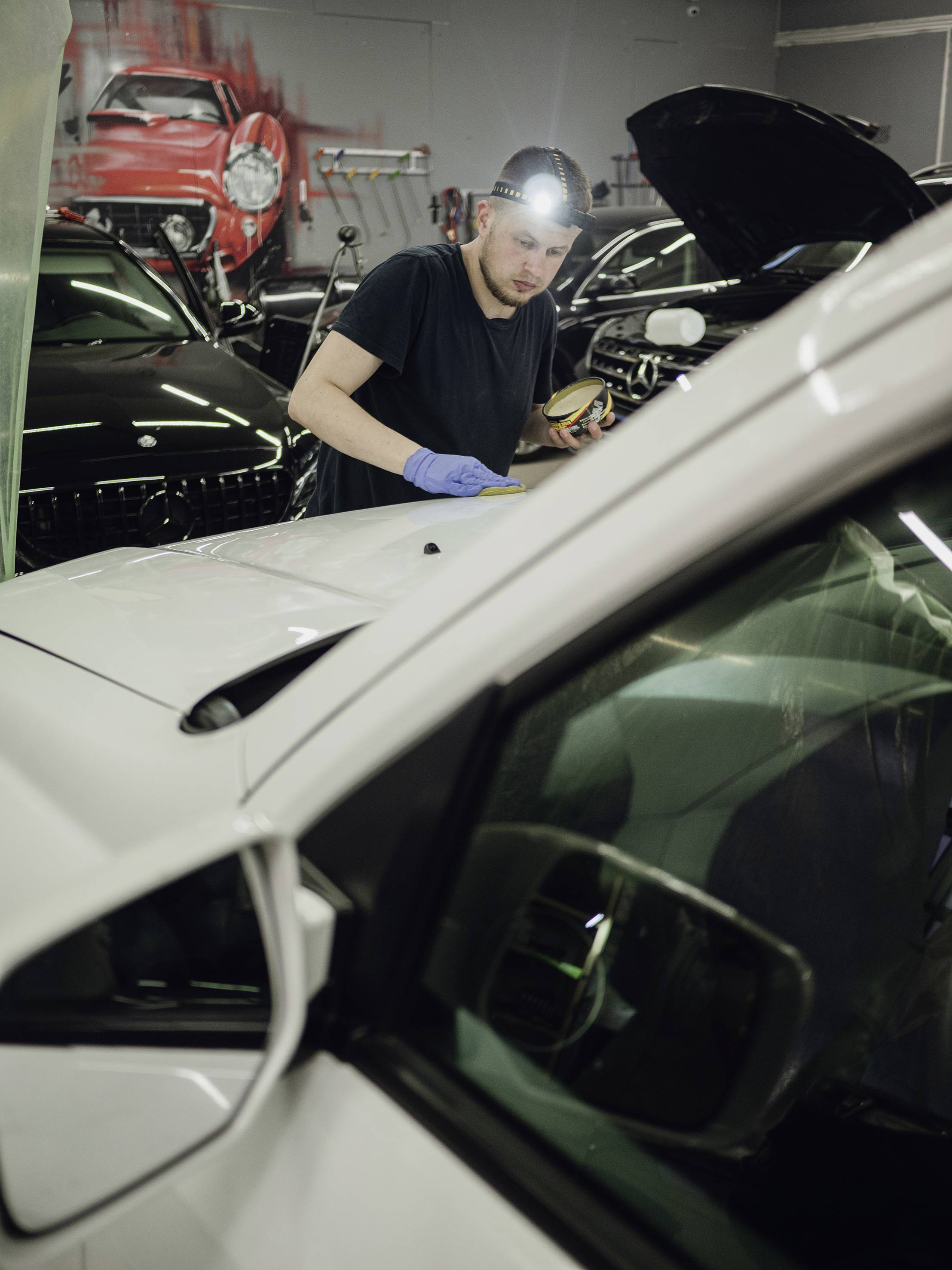 Ein Mechaniker mit Stirnlampe arbeitet in einer Werkstatt an einem Auto. Mehrere Autos sind im Hintergrund zu sehen, einige mit geöffneten Motorhauben.