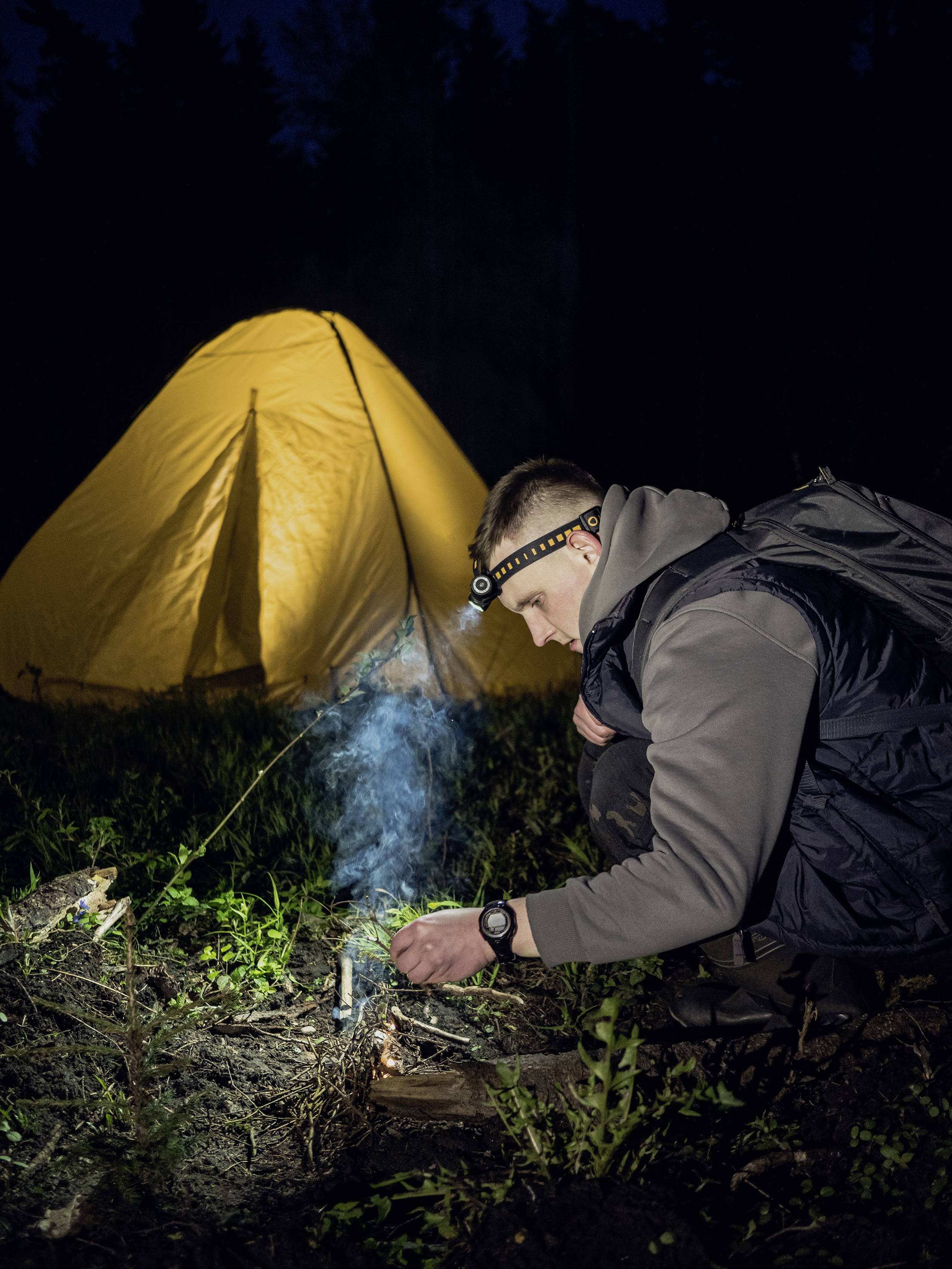 Eine Person entzündet ein kleines Feuer vor einem gelben Zelt bei Nacht. Die Umgebung ist dunkel, beleuchtet vom Zeltlicht.