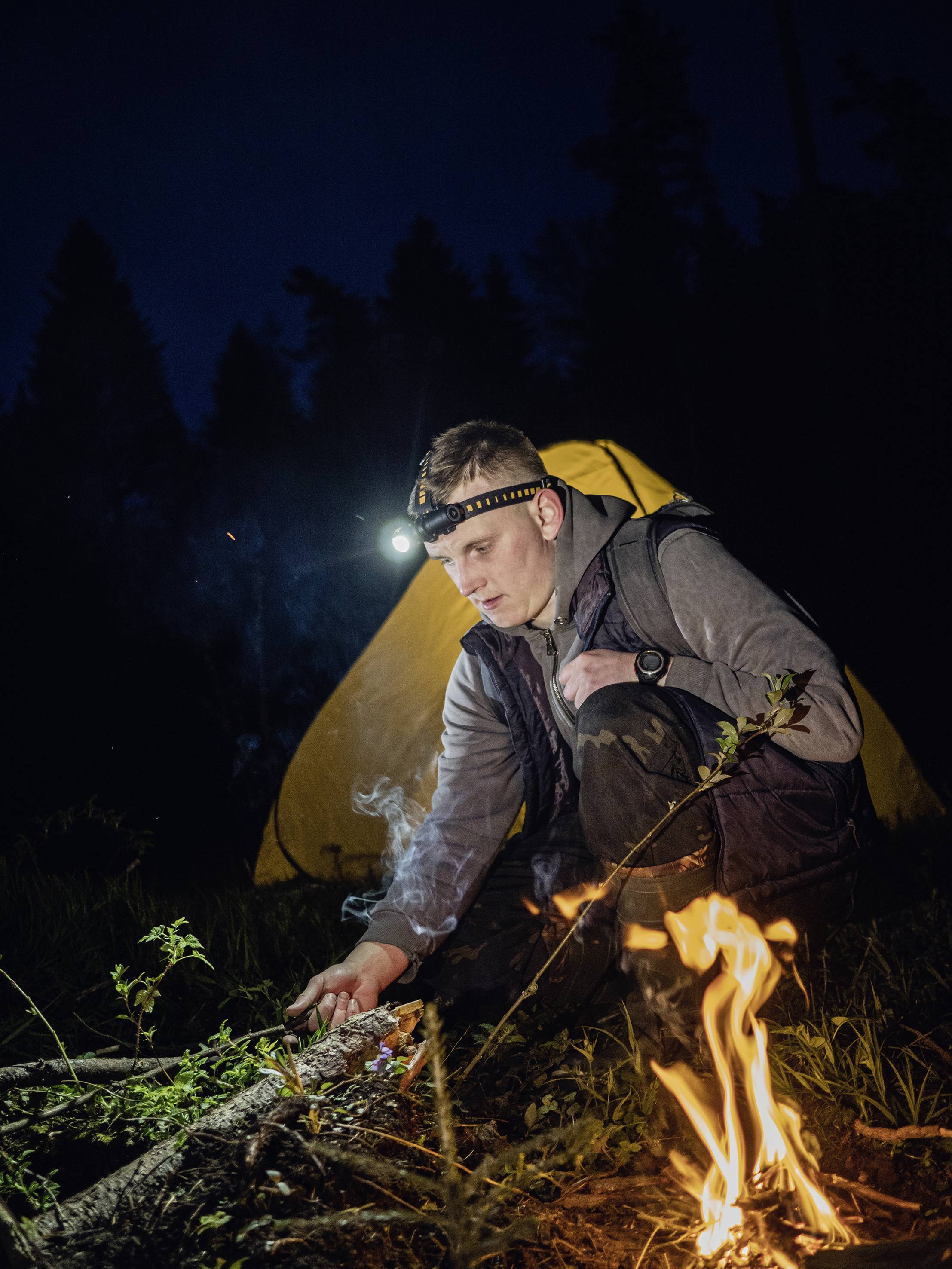 Ein Mann im Wald zündet abends ein Lagerfeuer an, während er eine Stirnlampe trägt. Ein gelbes Zelt steht im Hintergrund.