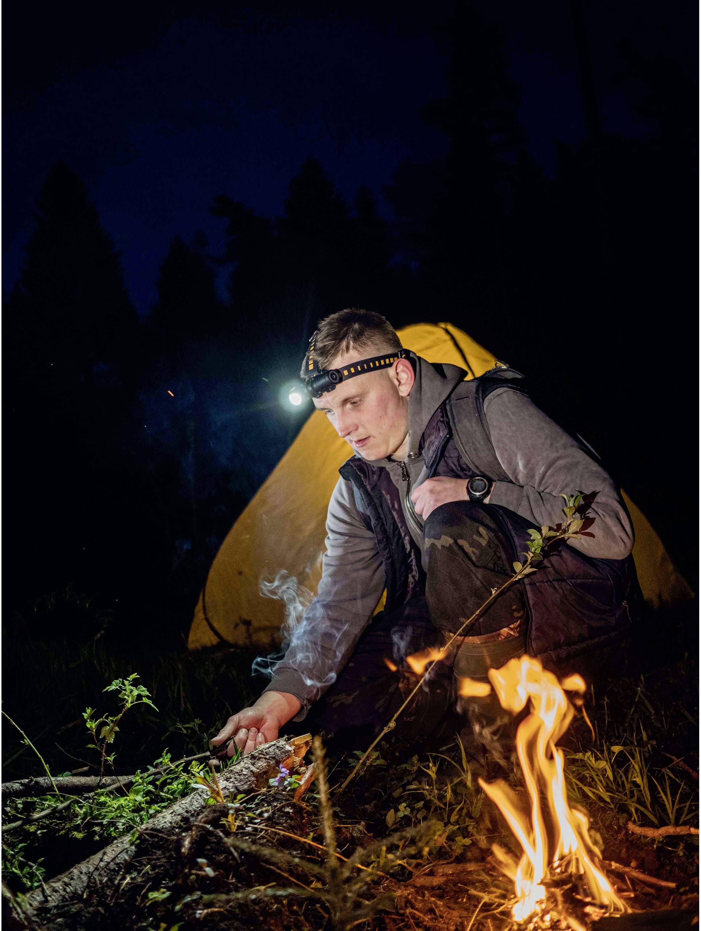 Ein Mann mit Stirnlampe hockt vor einem Zelt und entzündet ein kleines Lagerfeuer im Wald bei Nacht.