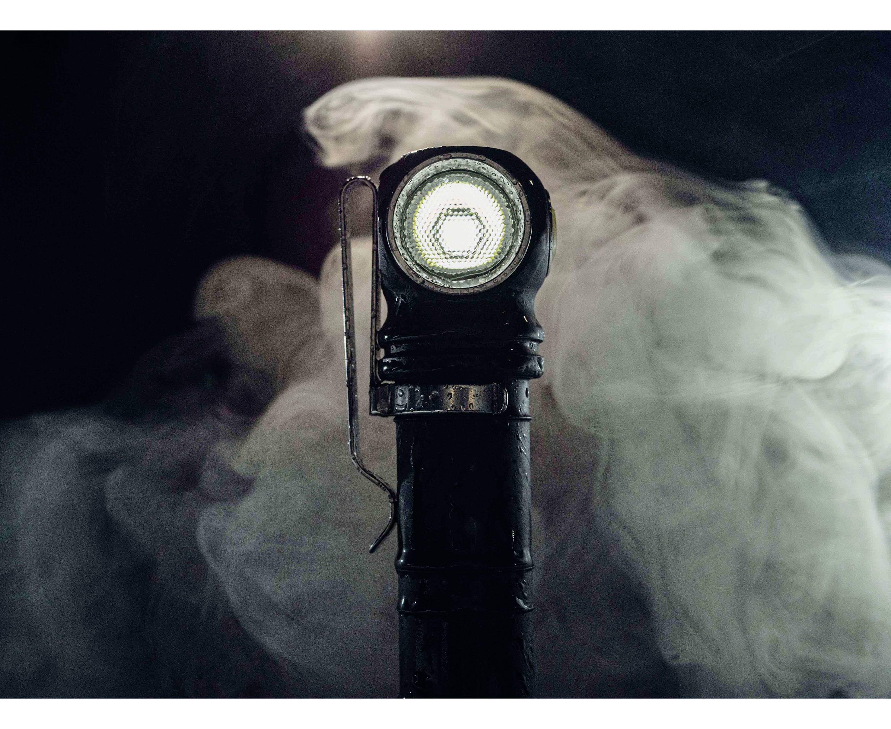 'Nahaufnahme einer alten Taschenlampe mit eingeschaltetem Licht, umgeben von aufsteigendem Rauch. Dunkler Hintergrund hebt das Licht hervor.'