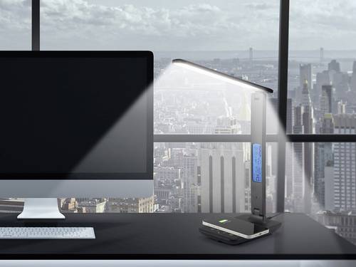 Moderne Bürolampe mit Display beleuchtet Schreibtisch und Monitor vor Stadtkulisse.