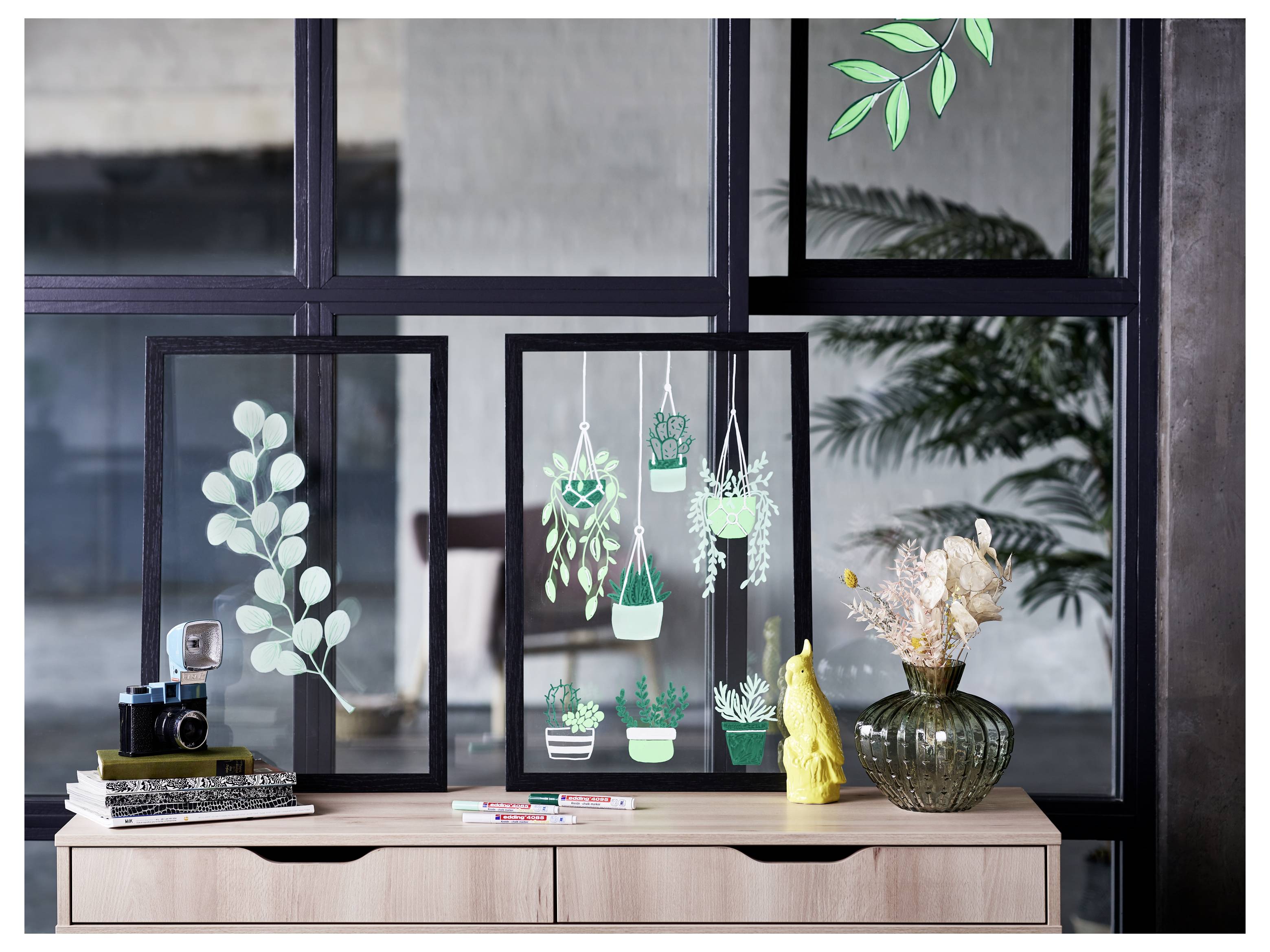 Dekorative Ausstellung von gerahmten Pflanzenkunstwerken auf einem Schrank, mit Büchern, einer Kamera, Stiften, einer goldenen Figur und einer Vase mit getrockneten Blumen vor einem Fenster.