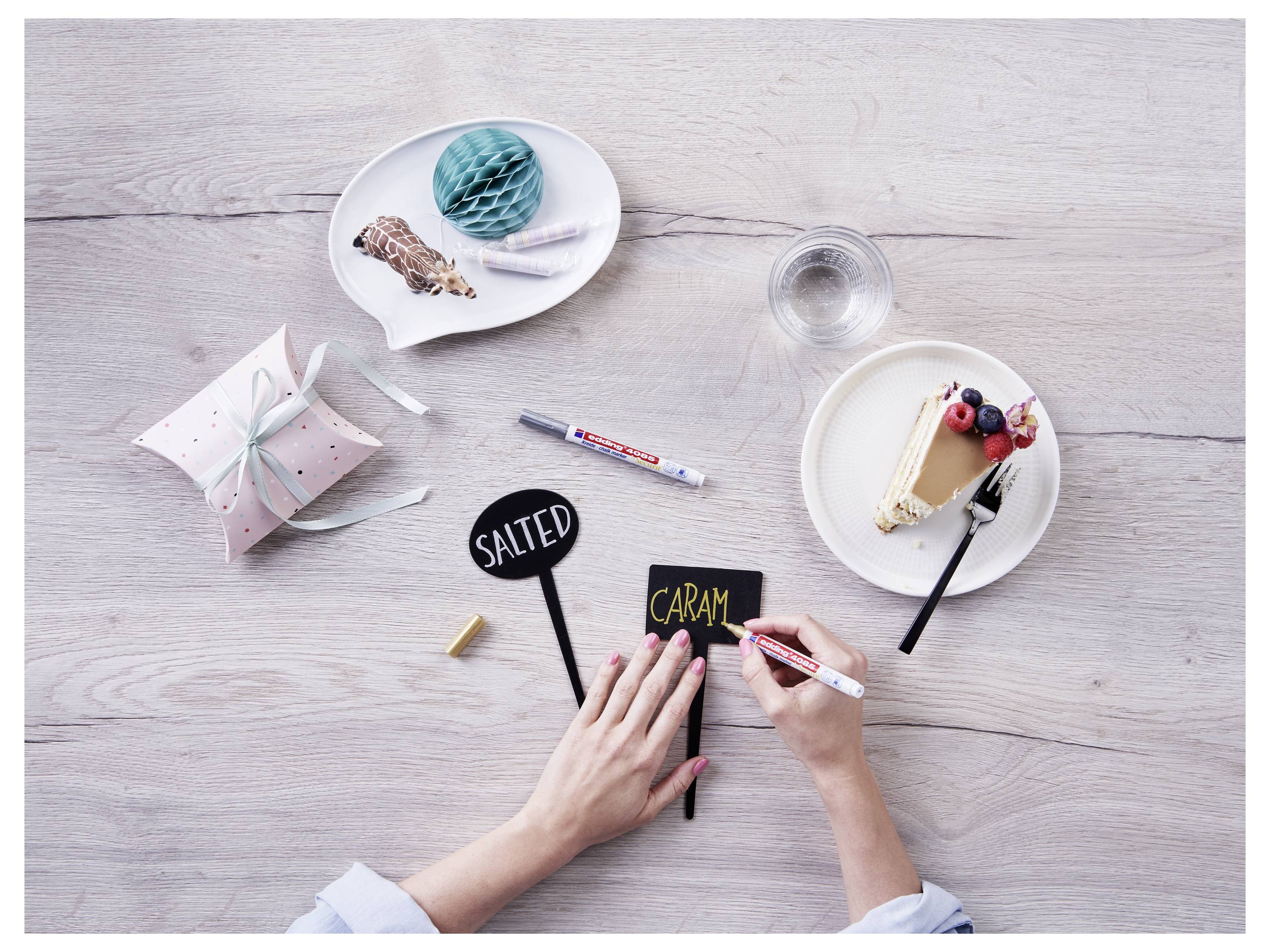 Hände schreiben "CARAM" auf ein kleines schwarzes Schild mit handwerklichen Desserts und Materialien, die auf einem hellen Holztisch angeordnet sind.