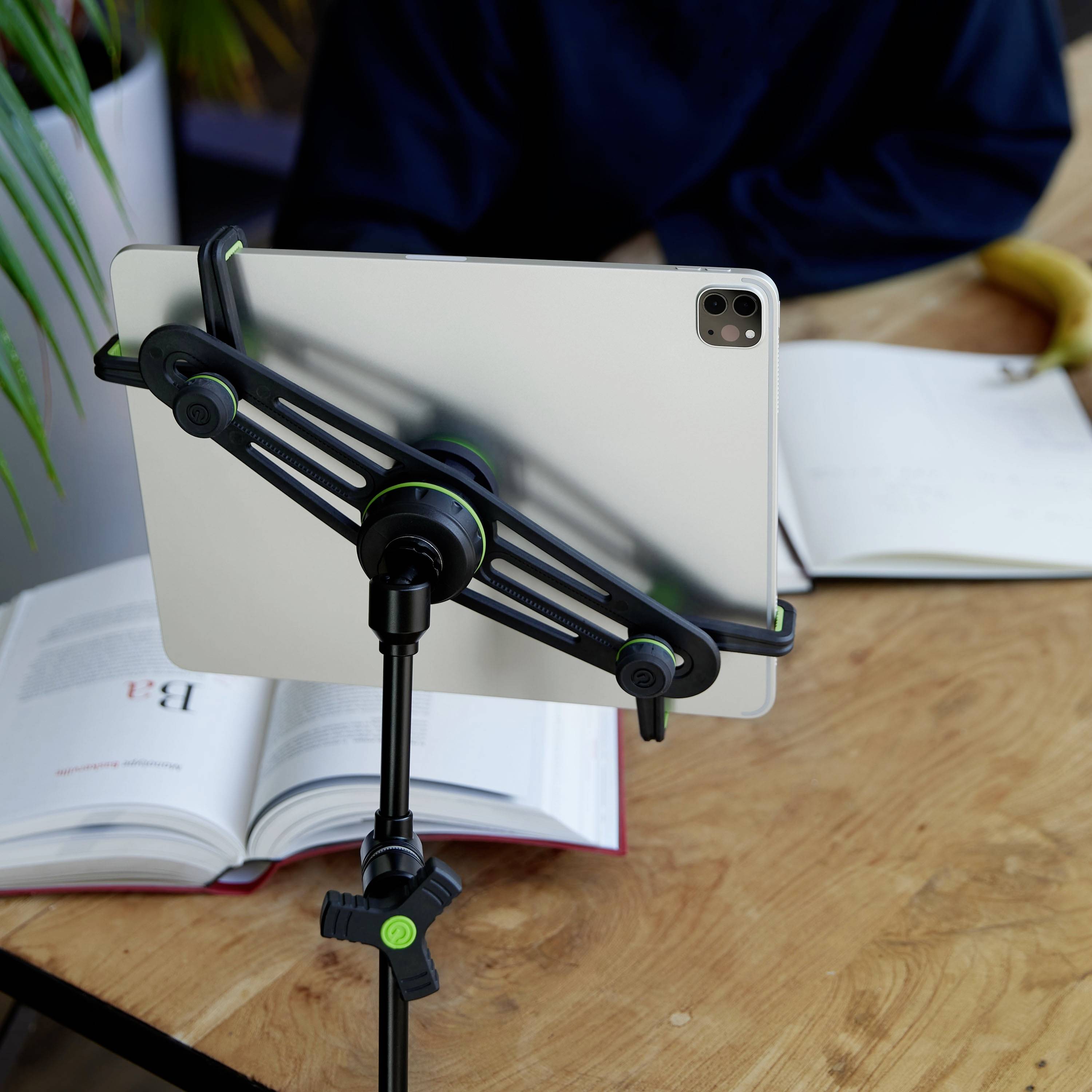 Ein Tablet auf einem Ständer steht auf einem Tisch zwischen aufgeschlagenen Büchern und einer Banane. Szenario: Arbeitsumgebung.