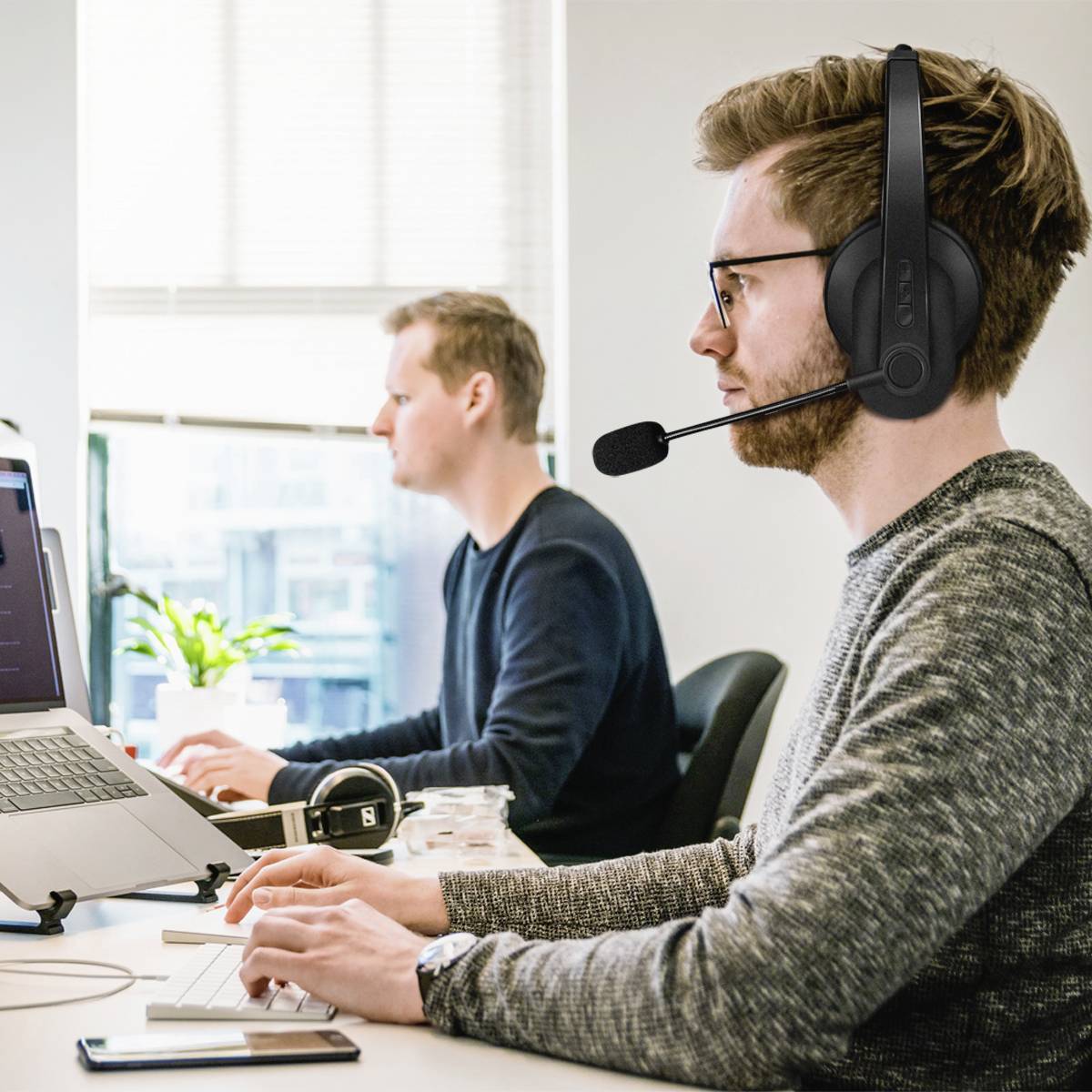 Zwei Männer arbeiten an Computern in einem Büro. Der vordere Mann trägt ein Headset mit Mikrofon. Im Hintergrund ist eine Pflanze am Fenster.