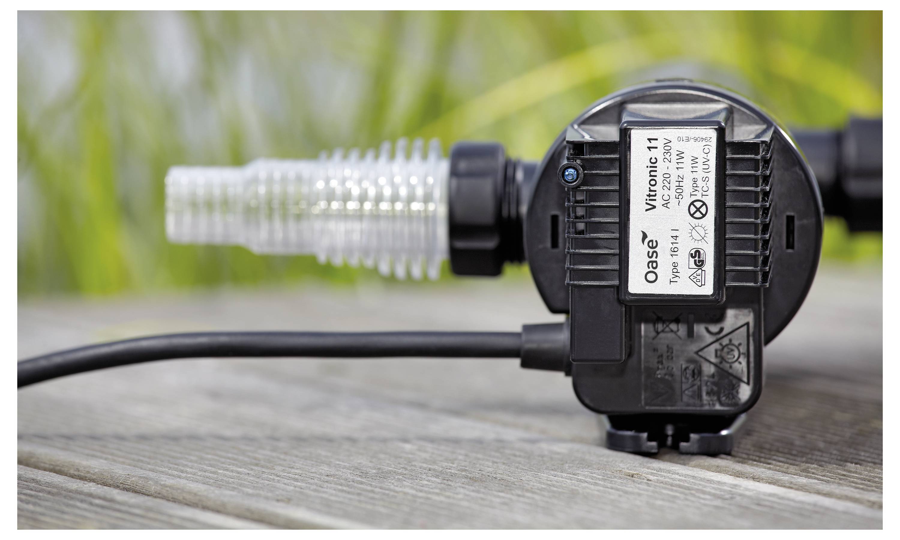 Eine kleine, schwarze Wasserpumpe mit der Aufschrift „Oase Vitronic 11" liegt auf einer Holzoberfläche und ist mit einem transparenten Spiralschlauch verbunden. Im Hintergrund befindet sich Grünbewuchs.