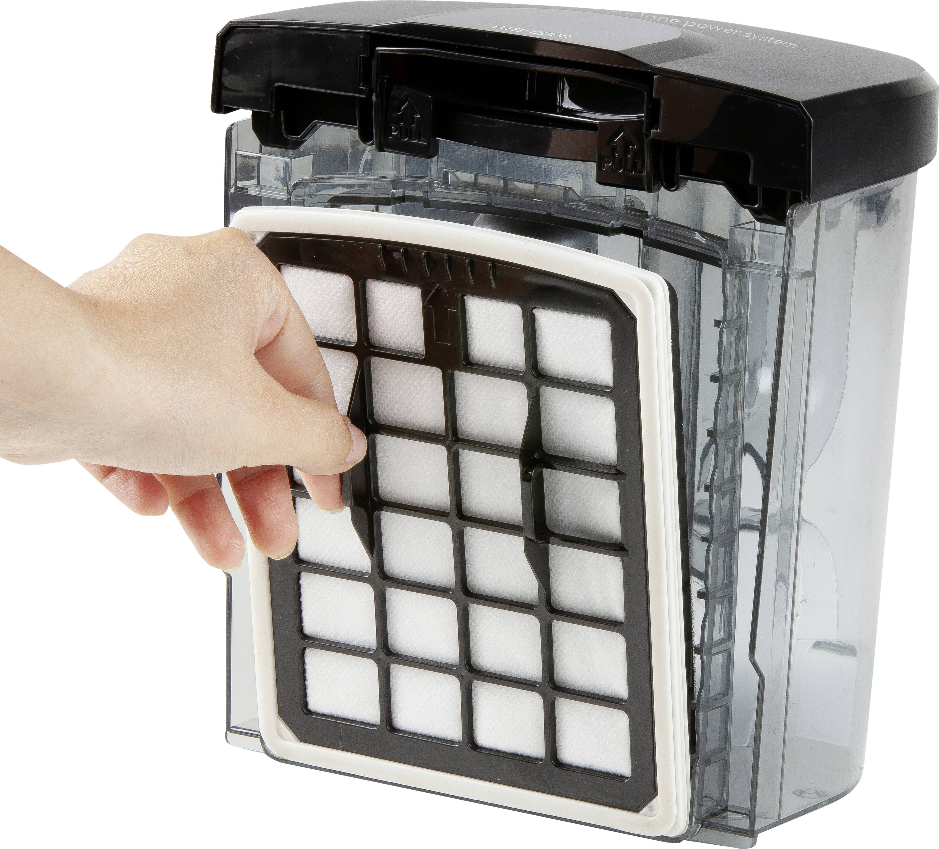 Eine Hand zieht den Filter aus einem rechteckigen, transparenten Staubsaugerbehälter.