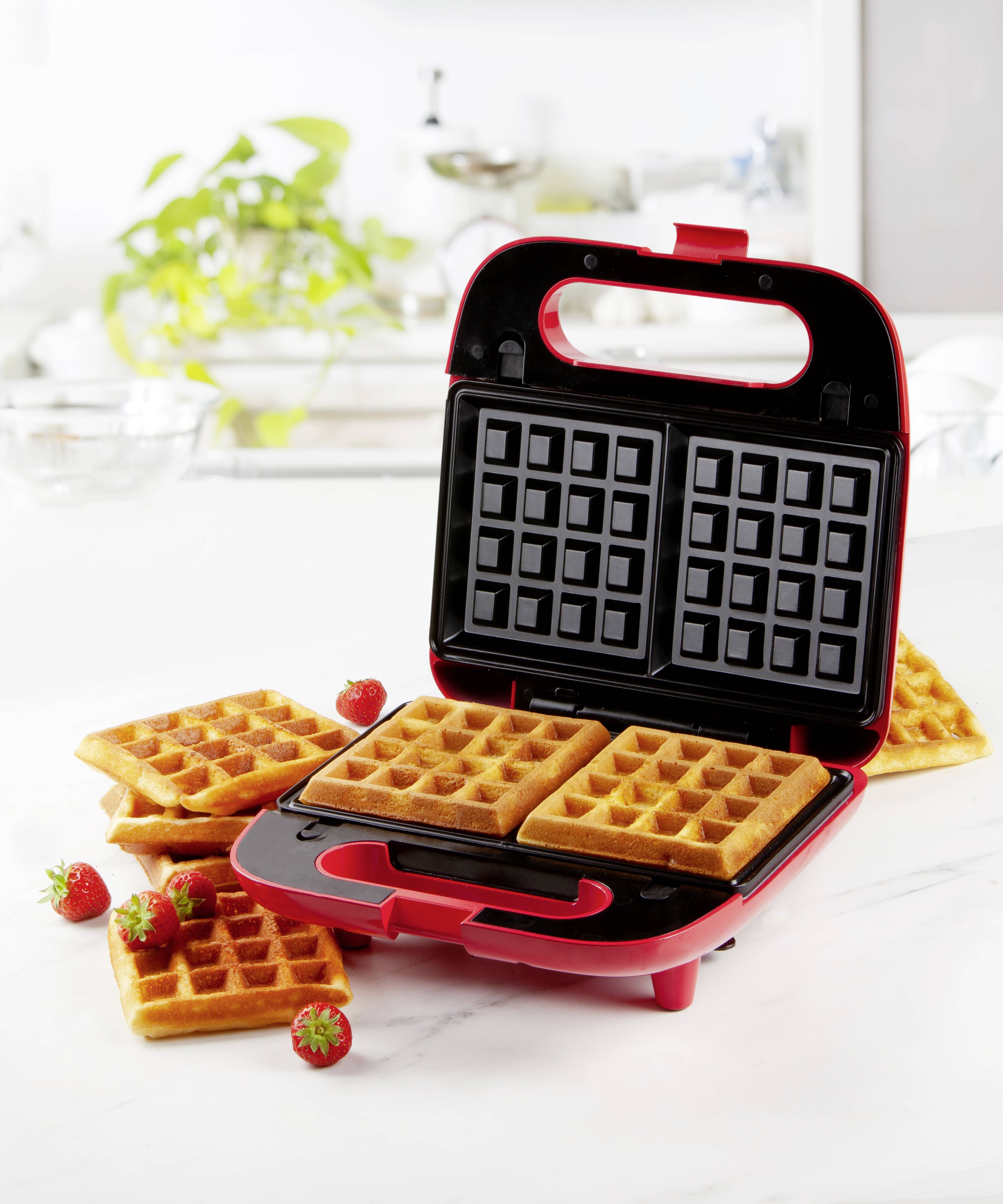 Ein rotes Waffeleisen mit zwei gebackenen Waffeln darin, umgeben von fertigen Waffeln und Erdbeeren auf einer weißen Küchenplatte.