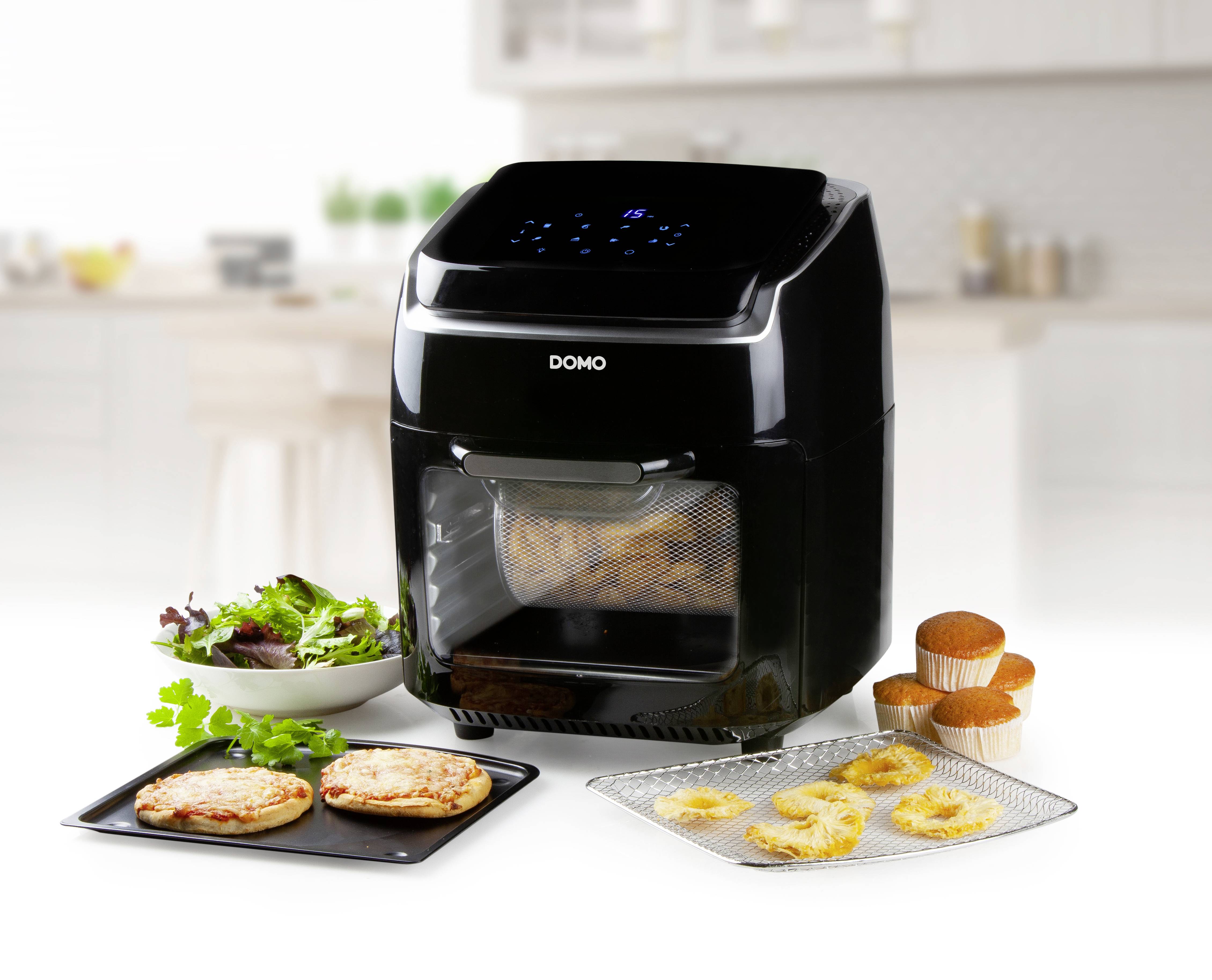 Ein schwarzer Airfryer steht auf einer Küchentheke, umgeben von einer Salatschüssel, zwei fertigen Pizzen und Muffins.